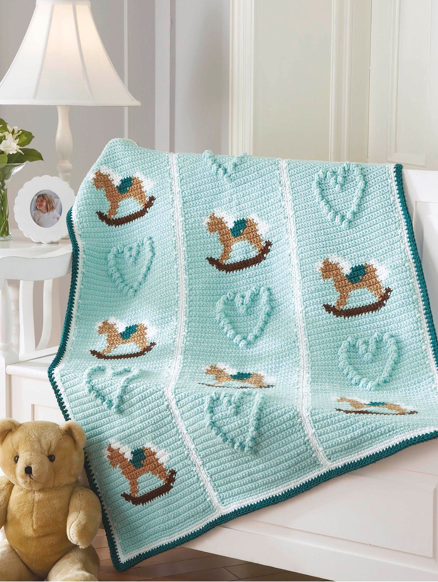 The Leisure Arts Crochet Bedtime Blankies features a light blue baby blanket with brown rocking horse and heart designs, draped over a white chair beside a beige teddy bear and photo frame—ideal for cozy bedtime moments.