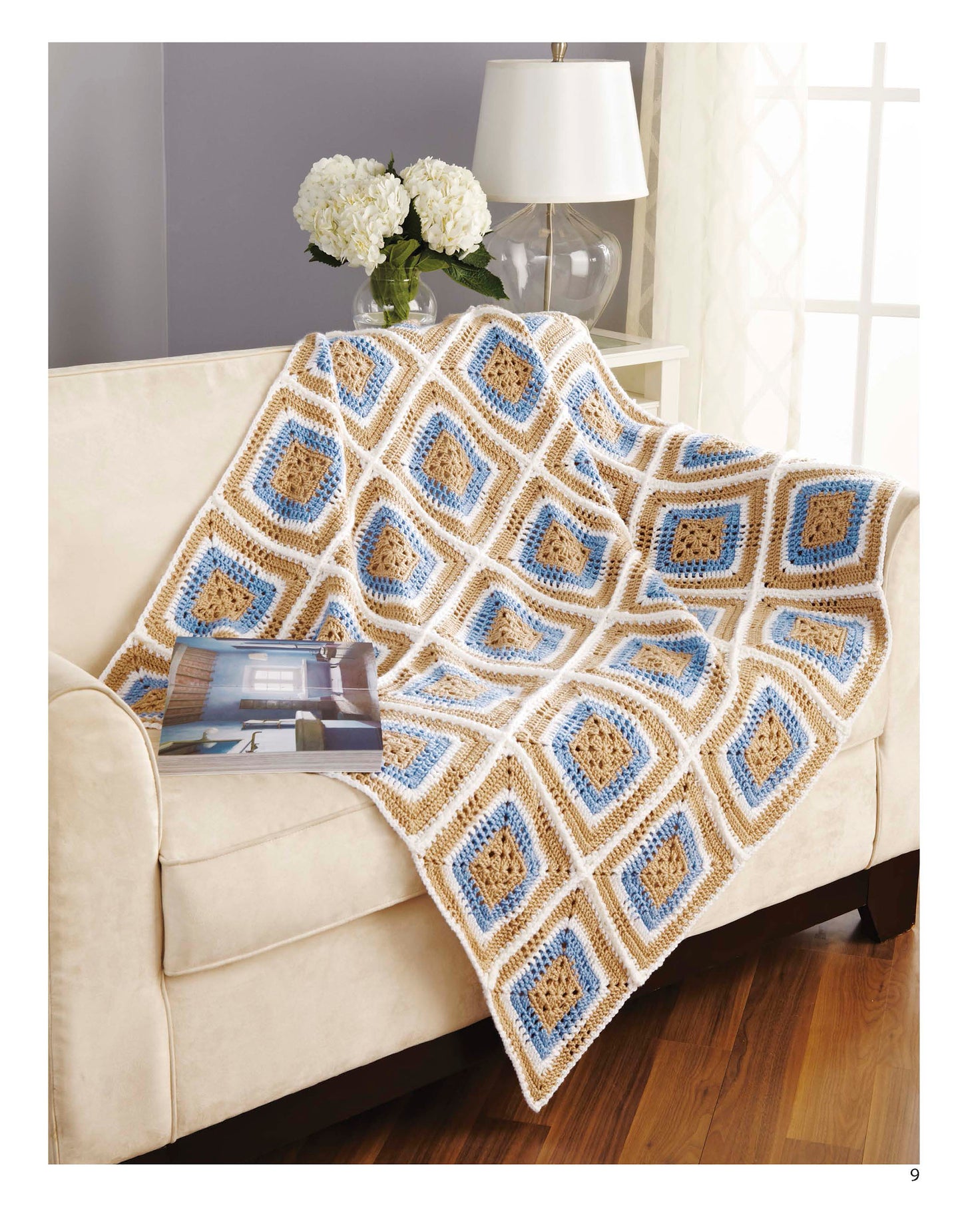 A cream-colored couch features the Cozy Comfort Afghans by Leisure Arts—a blue, beige, and white diamond-patterned crocheted blanket with a magazine atop. Nearby are a lamp, white flowers, and sunlight streaming through the window.