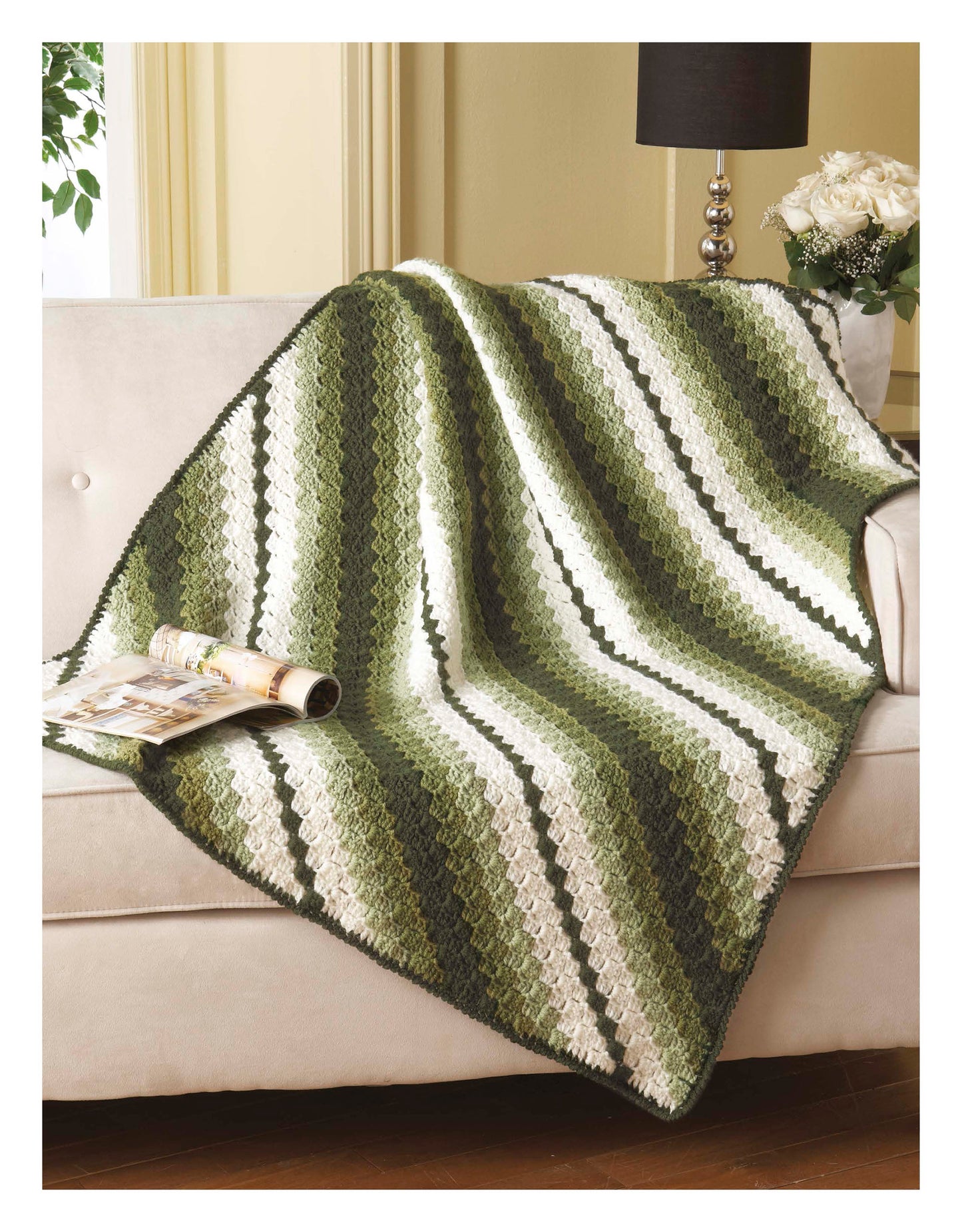 The Cozy Comfort Afghans by Leisure Arts, featuring green, white, and olive stripes in worsted weight yarn, is draped over a light sofa with a magazine on top. A black lamp and white flowers sit on a nearby table.