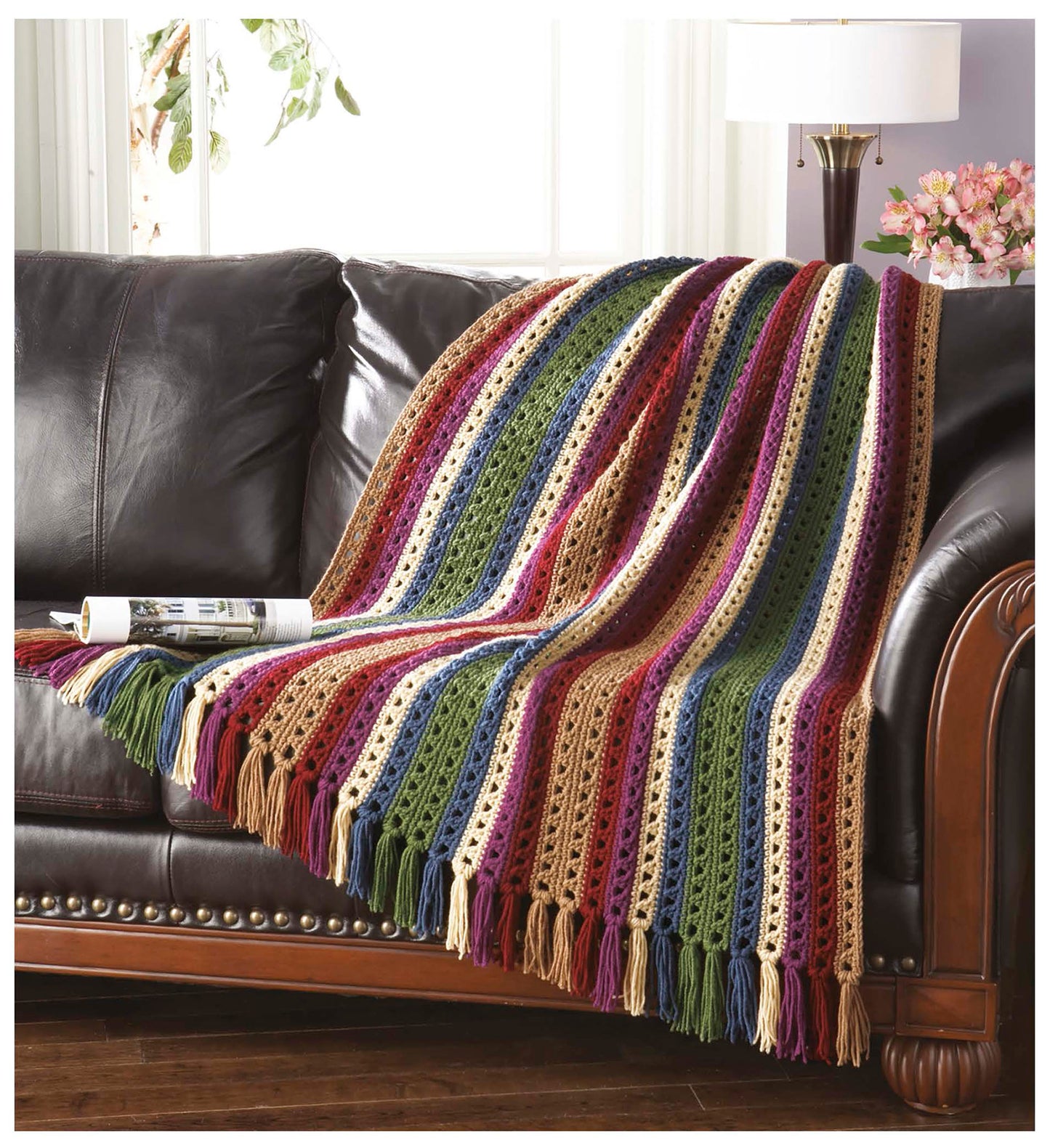 The Cozy Comfort Afghans by Leisure Arts, crafted from worsted weight yarn in vibrant stripes with tassels, is draped over a dark leather sofa. A nearby magazine and a floral lamp on the side table complete the cozy scene.