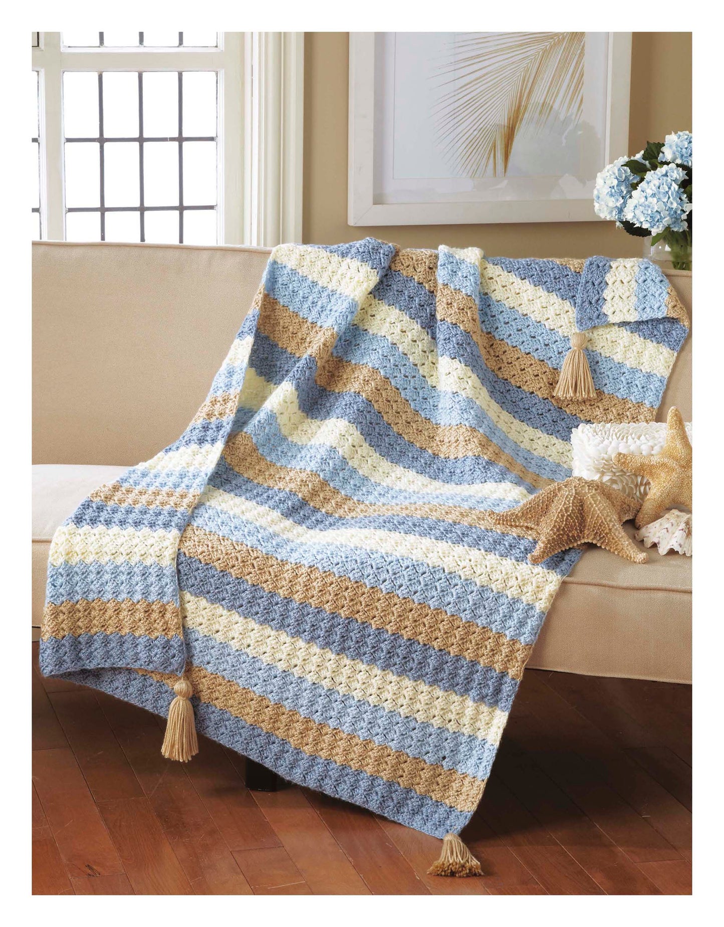 The Cozy Comfort Afghans by Leisure Arts—a striped, tasseled blanket in blue, beige, and cream—crafted with worsted weight yarn, is draped over a beige sofa near a starfish and a vase of blue flowers.