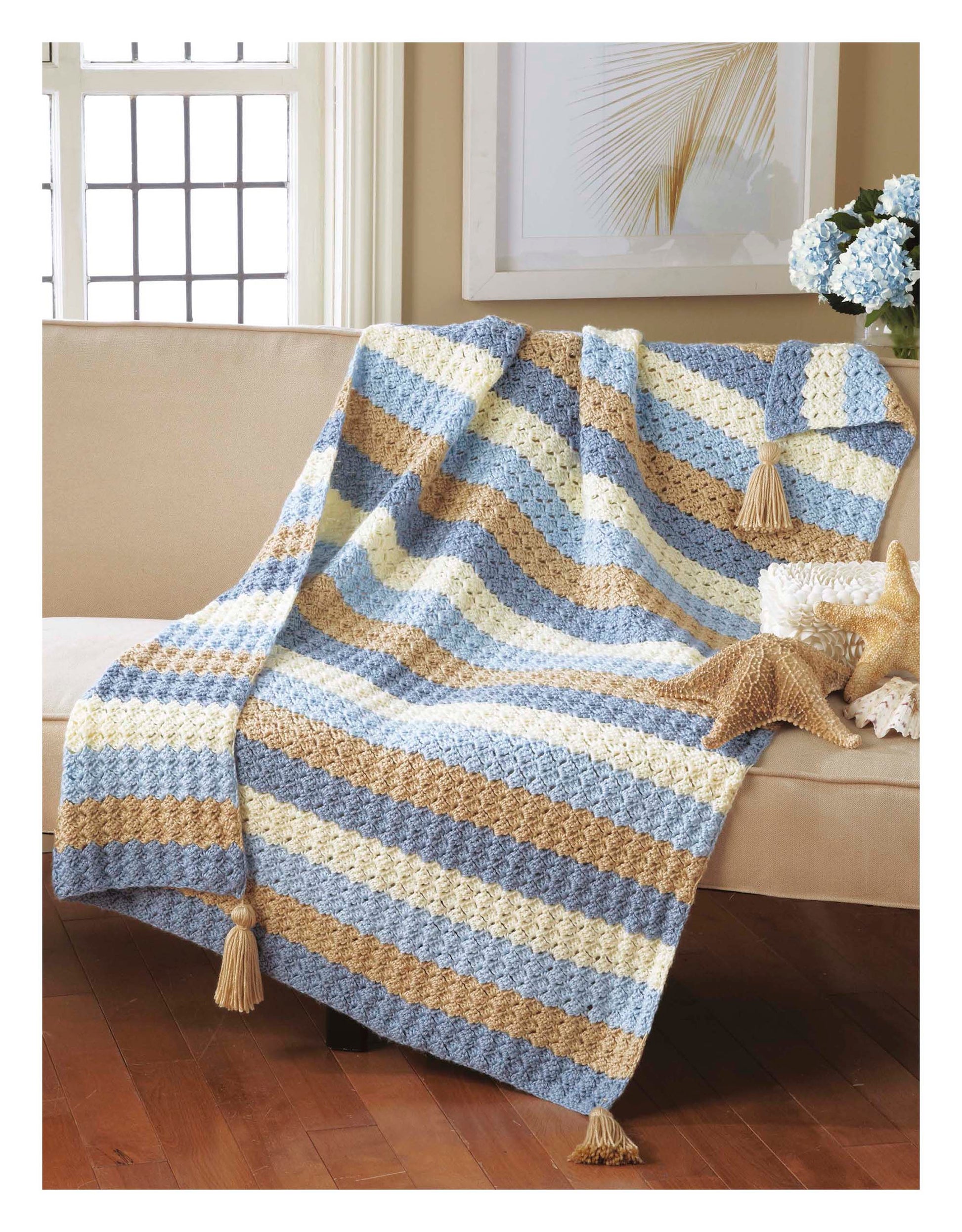 The Cozy Comfort Afghans by Leisure Arts—a striped, tasseled blanket in blue, beige, and cream—crafted with worsted weight yarn, is draped over a beige sofa near a starfish and a vase of blue flowers.