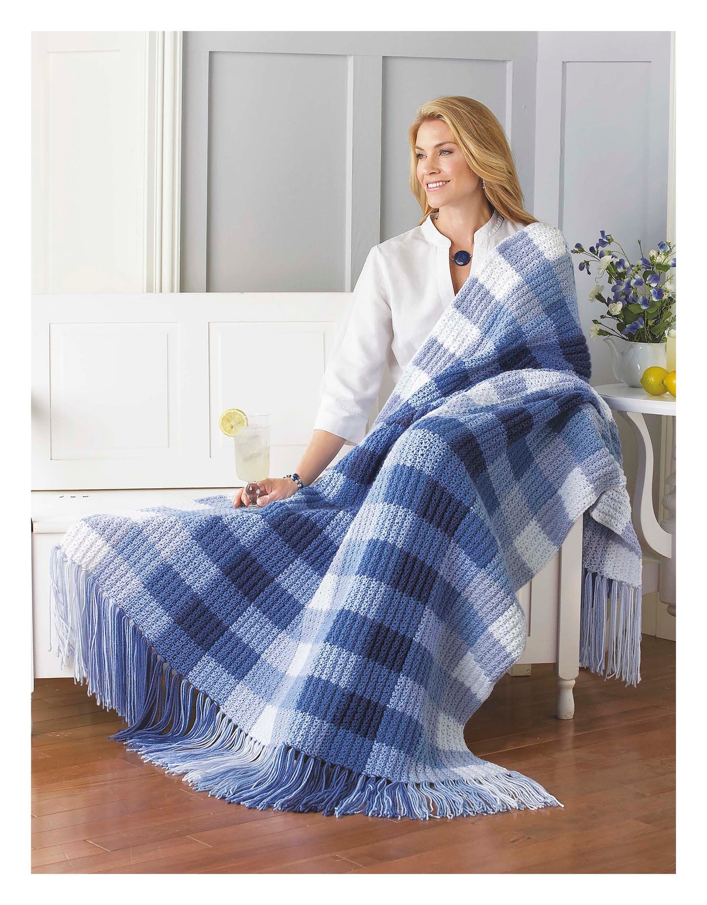 A smiling woman sits on a white bench wrapped in a Cozy Comfort Afghans blanket from Leisure Arts, featuring blue and white plaid with classic designs, as a glass of lemonade with lemons rests on the table beside her.