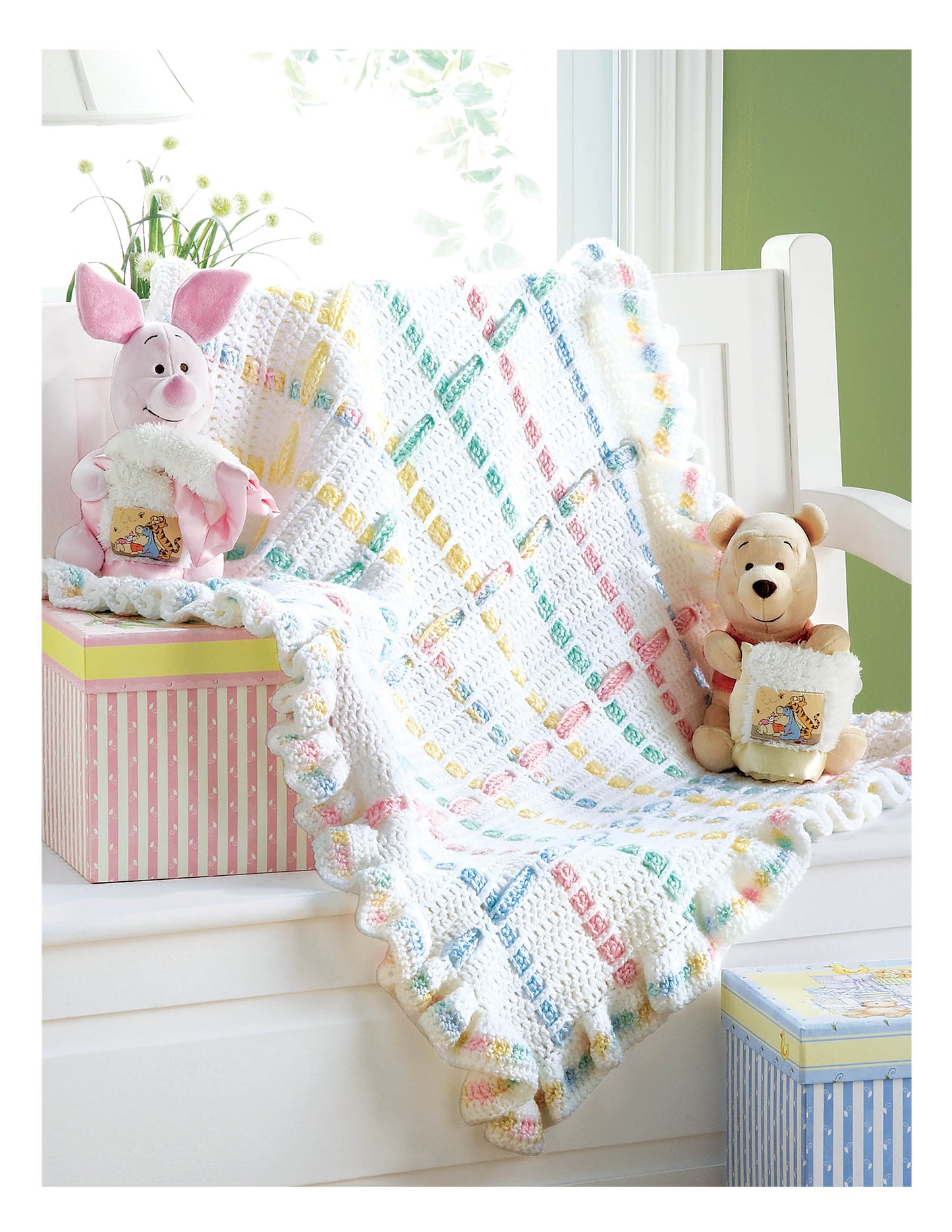 A Leisure Arts Cuddle Me Baby Afghans pastel crochet blanket drapes over a white bench, with Piglet and Pooh plush toys on striped boxes holding matching blankets—a bright, cozy scene perfect as a baby shower gift.