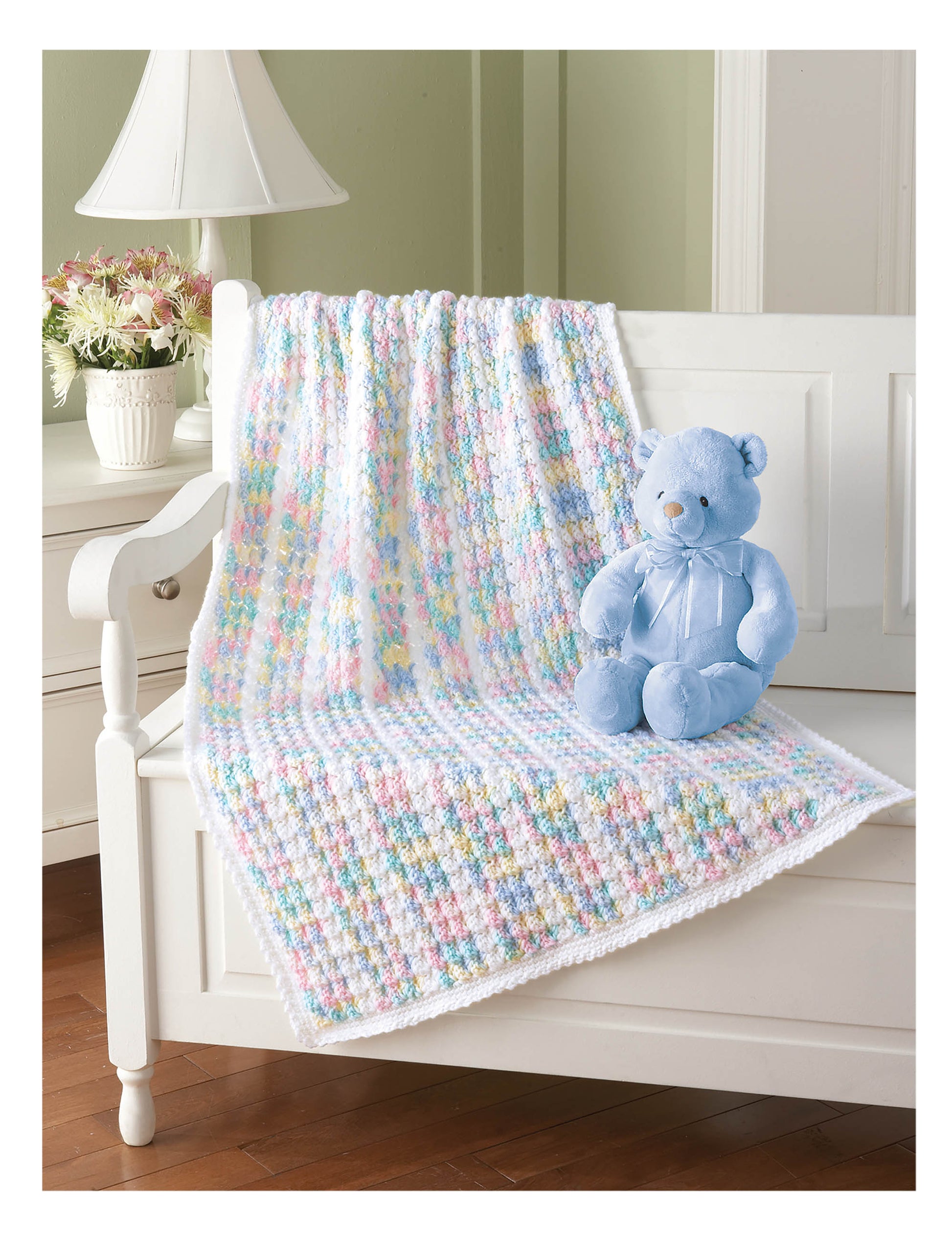 A pastel-colored crocheted baby blanket from the Leisure Arts Cuddle Me Baby Afghans collection is draped over a white bench beside a plush blue teddy bear and a vase of flowers, creating a charming baby shower gift scene.