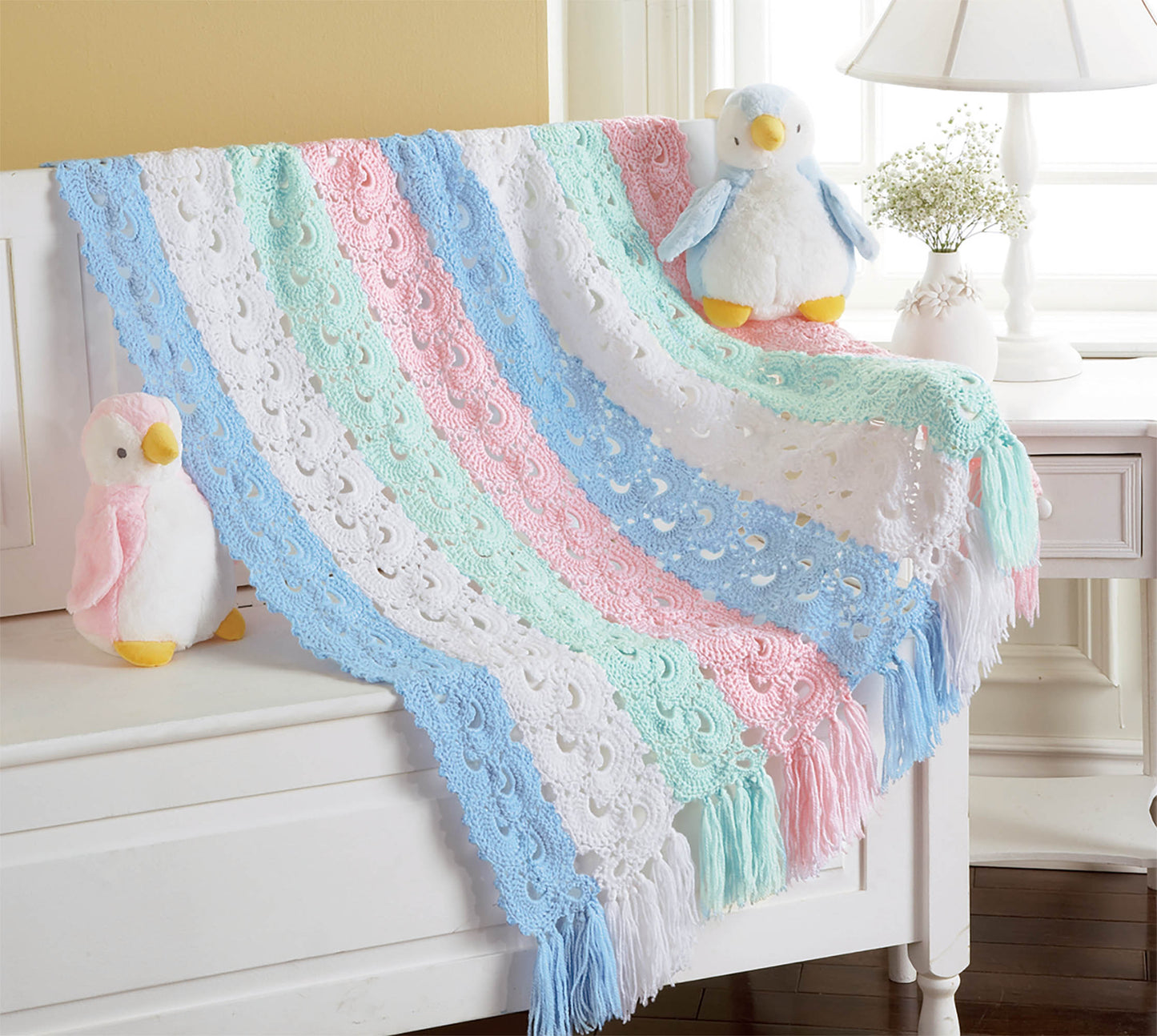 The "Cuddle Me Baby Afghans" by Leisure Arts, featuring pastel blue, pink, green, and white stripes from intermediate patterns, is draped over a bench with plush penguin toys—an adorable baby shower gift idea.