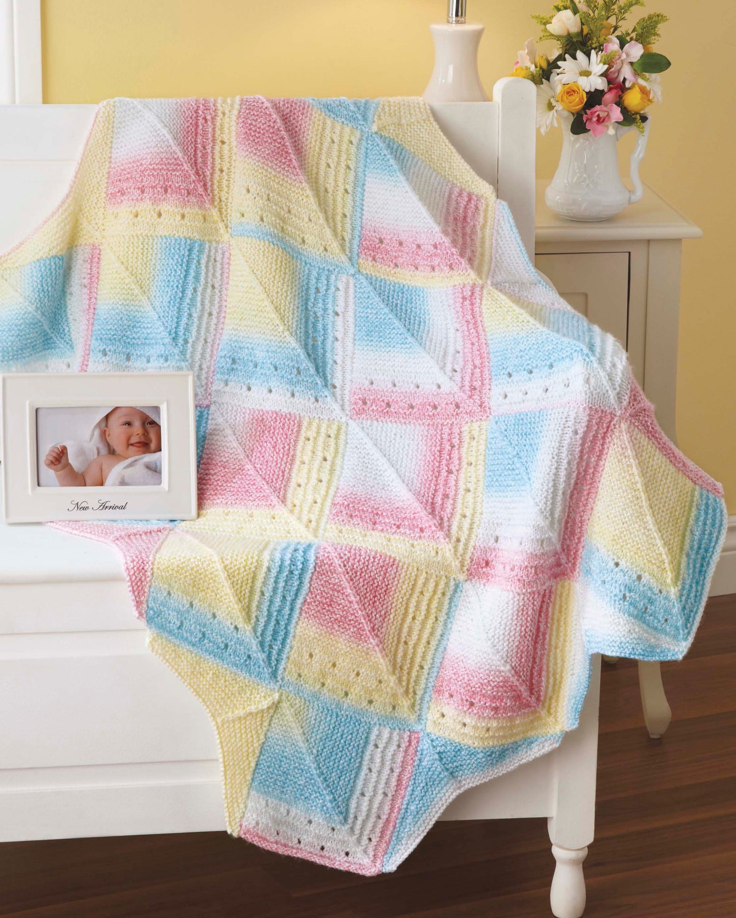 The Timeless Knit Baby Blankets by Leisure Arts, in soft pastels and various yarn weights, drape over a white crib. On a nearby white table sit a framed baby photo and a vase of flowers.