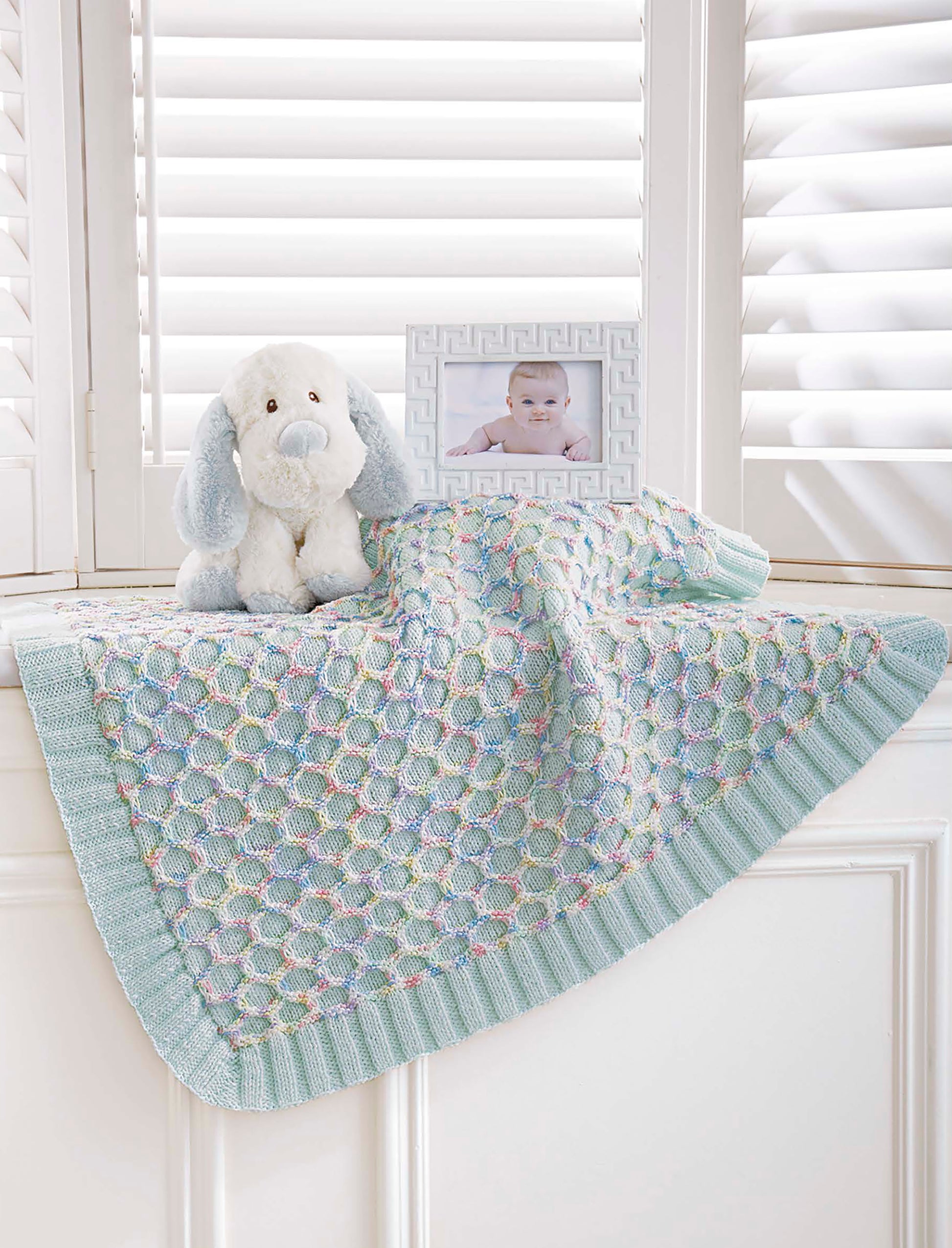 The Timeless Knit Baby Blanket by Leisure Arts, featuring a textured pastel design in various yarn weights, is draped over a windowsill beside a white and blue stuffed dog and a baby photo, with soft light streaming through white shutters.