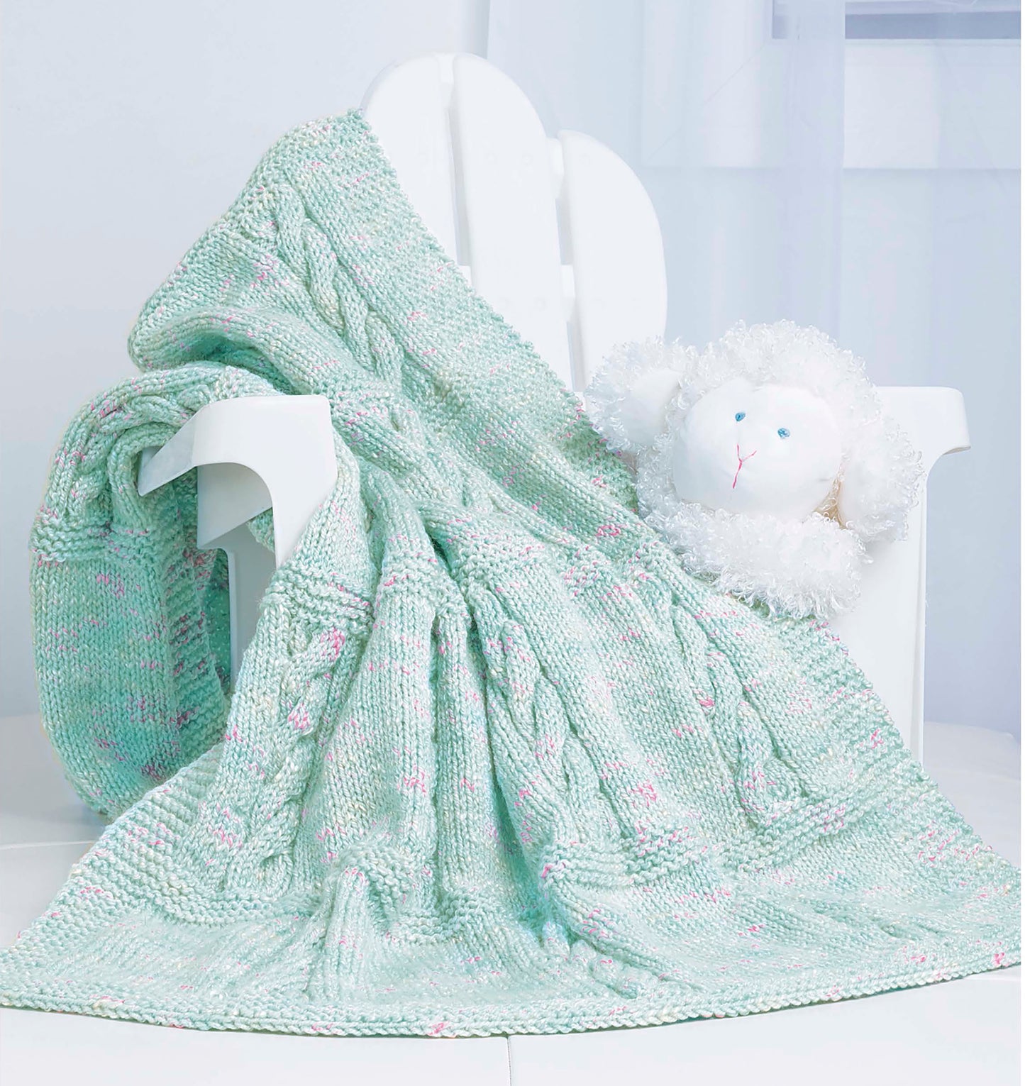 The Timeless Knit Baby Blanket by Leisure Arts, in pale green with pink accents, is draped over a white wooden chair beside a plush sheep toy in a bright, softly lit room.