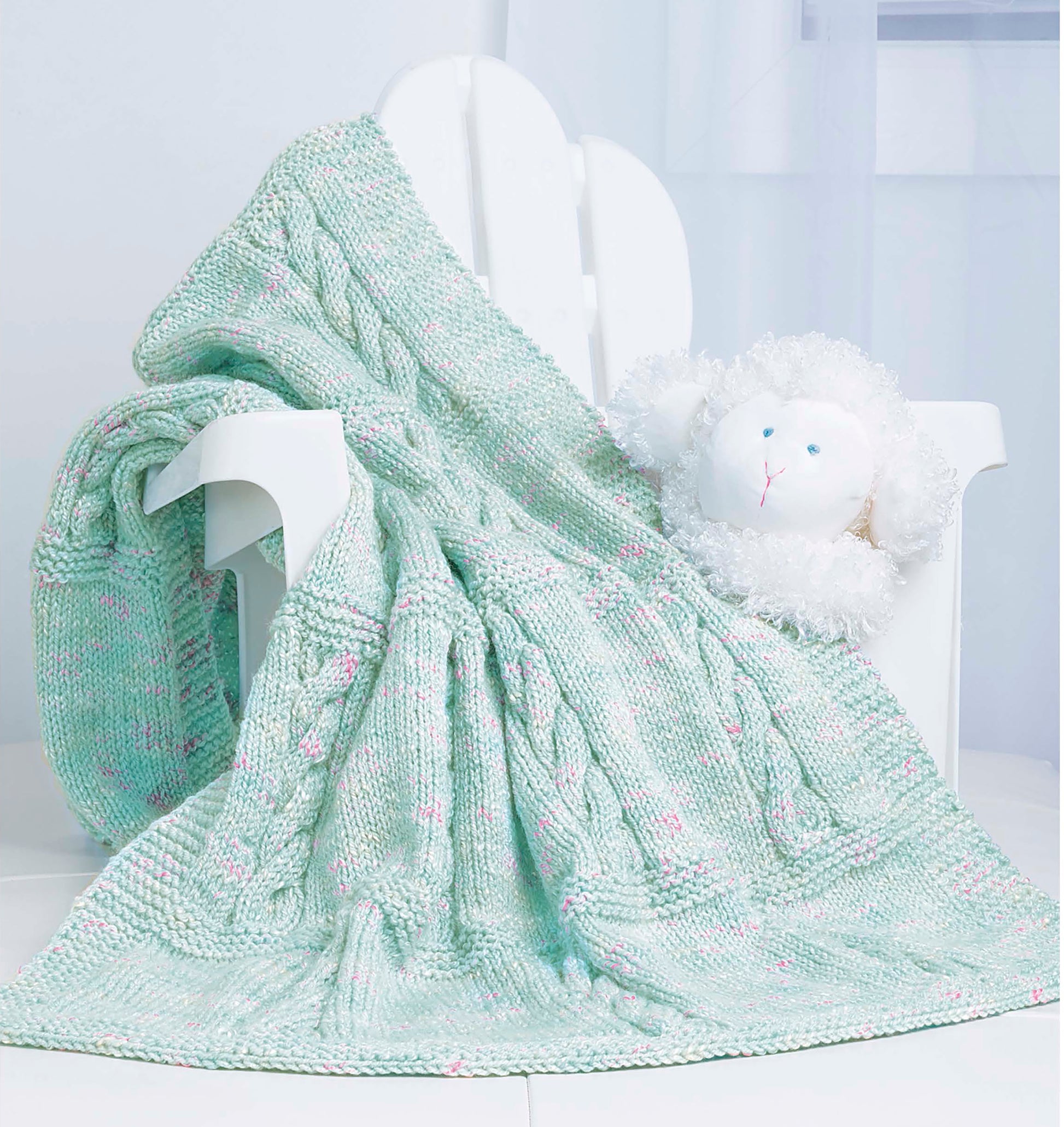 The Timeless Knit Baby Blanket by Leisure Arts, in pale green with pink accents, is draped over a white wooden chair beside a plush sheep toy in a bright, softly lit room.
