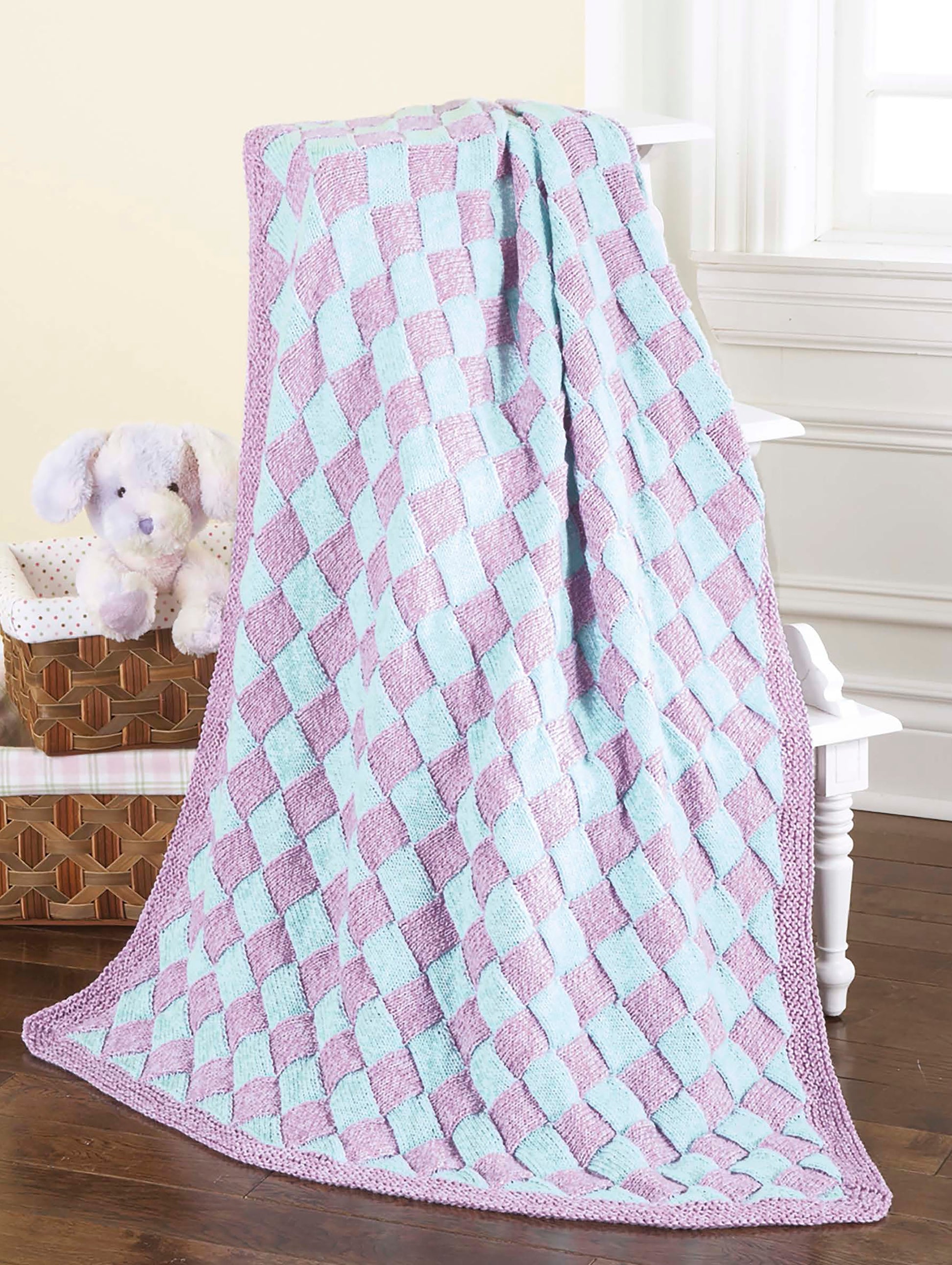 A Timeless Knit Baby Blanket by Leisure Arts in pastel blue and lavender is draped over a white chair near a basket holding a plush purple bunny and folded blankets, creating a cozy, sunlit atmosphere.