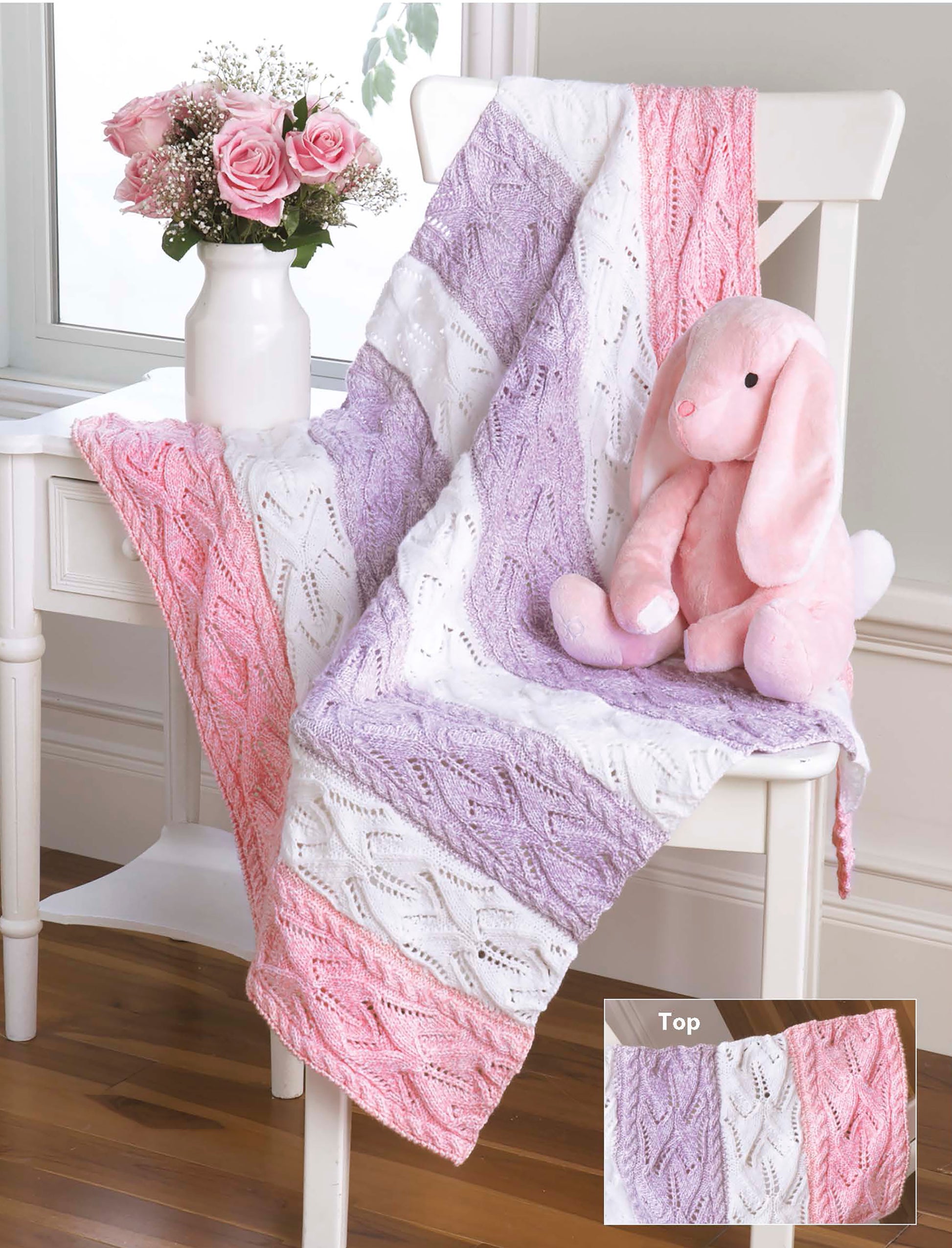 The Timeless Knit Baby Blanket by Leisure Arts in pink, purple, and white drapes over a white chair beside pink roses, with a plush bunny perched on the seat. An inset highlights the blanket’s detailed texture.