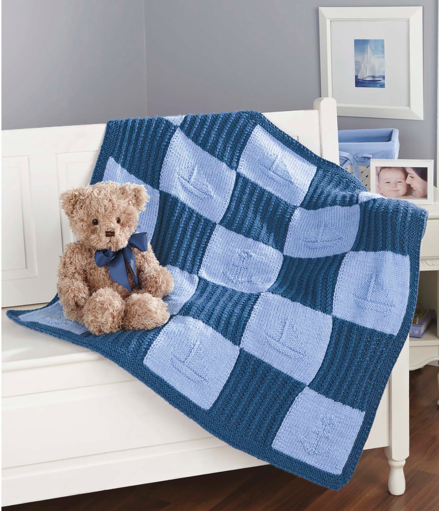 A plush teddy bear with a blue ribbon sits on a white bench draped with a blue patchwork sailboat blanket—just one of the charming projects in "Timeless Knit Baby Blankets" by Leisure Arts, ideal for intermediate knitters using various yarn weights.