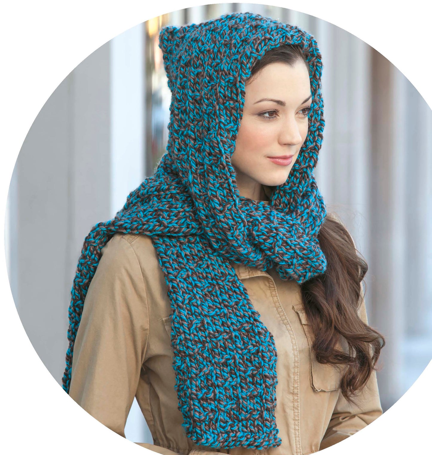Wearing a tan coat and a chunky, blue-and-gray knit scarf with a hood, a woman stands outside and smiles—perfect inspiration for projects from the Quick Knit Neckwear Digital Download by Leisure Arts.