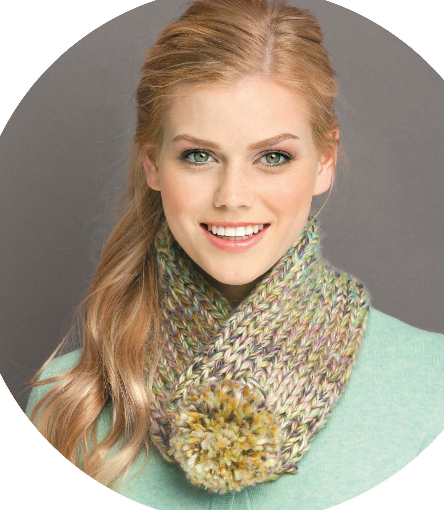 A smiling woman with long blonde hair models a chunky, multicolored knit scarf with pom-pom, paired with a mint green top against a gray background. Get inspired for scarves and cowls—find the pattern in Leisure Arts' Quick Knit Neckwear Digital Download.