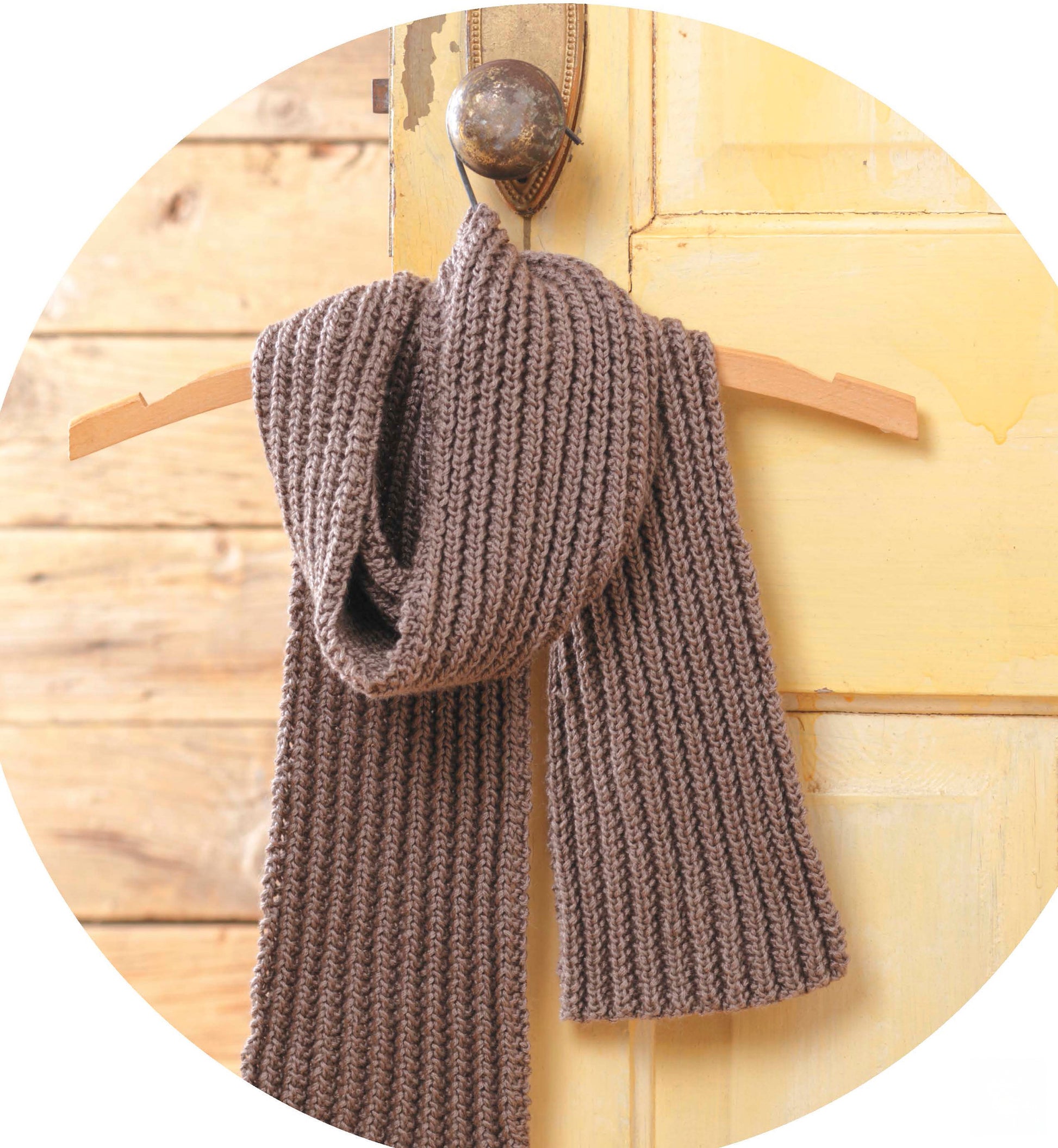 A chunky brown ribbed scarf is draped on a hanger against a rustic yellow door, offering cozy inspiration for beginner knitters. Create similar pieces with the Quick Knit Neckwear Digital Download by Leisure Arts.