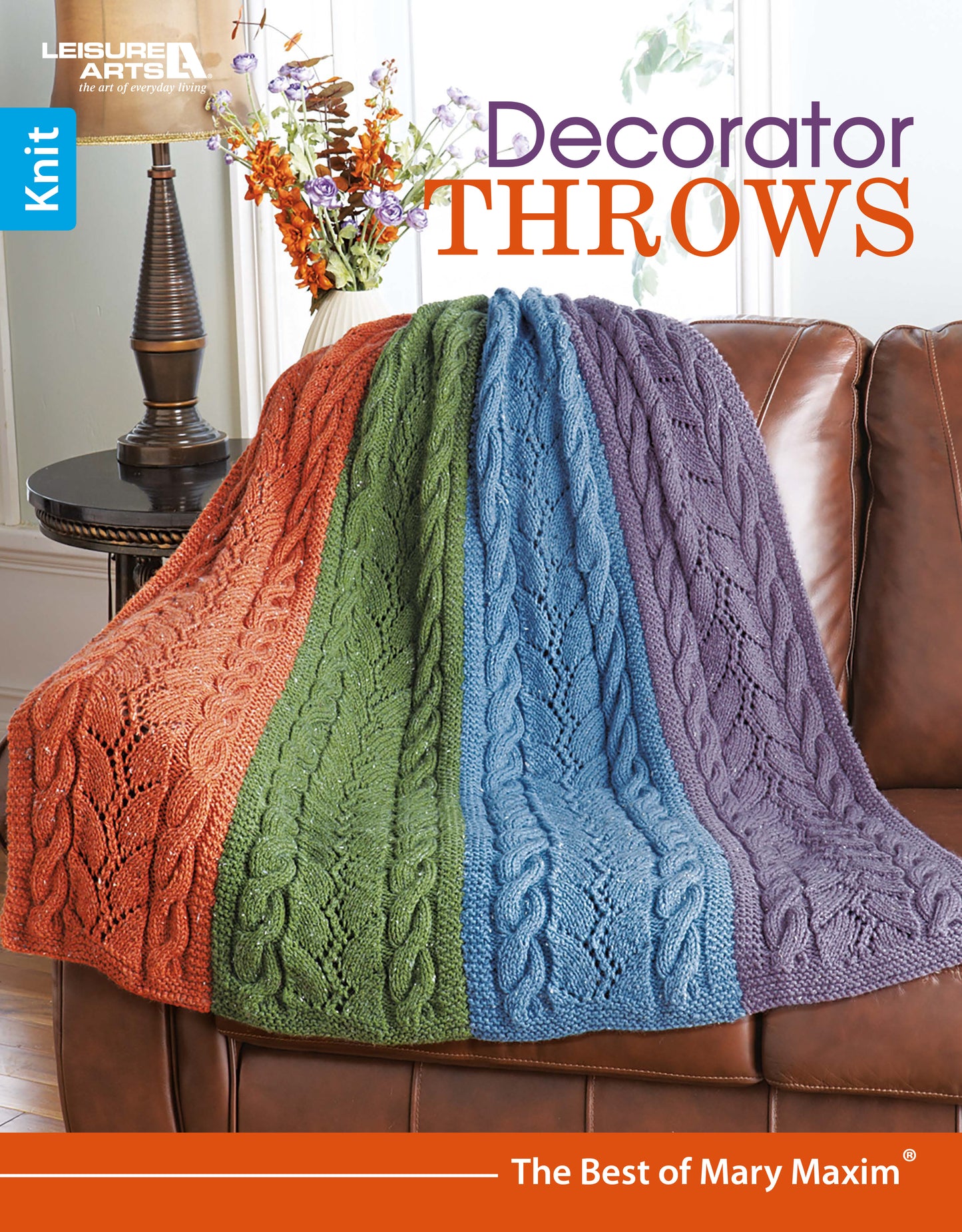 A Leisure Arts “Decorator Throws—The Best of Mary Maxim” multicolored knit throw in orange, green, blue, and purple is draped over a brown leather couch; flowers in a vase are on a table nearby.