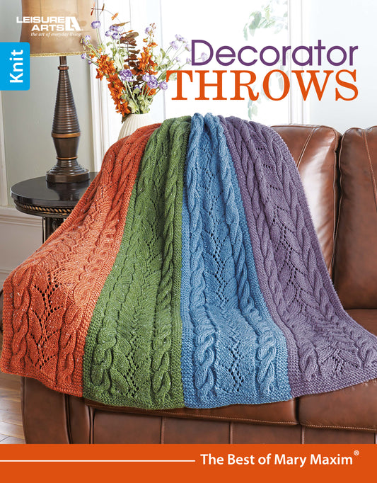 A Leisure Arts “Decorator Throws—The Best of Mary Maxim” multicolored knit throw in orange, green, blue, and purple is draped over a brown leather couch; flowers in a vase are on a table nearby.