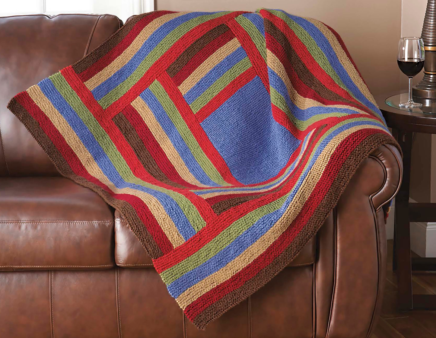 The Decorator Throws—The Best of Mary Maxim by Leisure Arts, a colorful knit blanket with blue, red, green, tan, and brown stripes, is draped over a brown leather sofa beside a side table holding a glass of red wine.