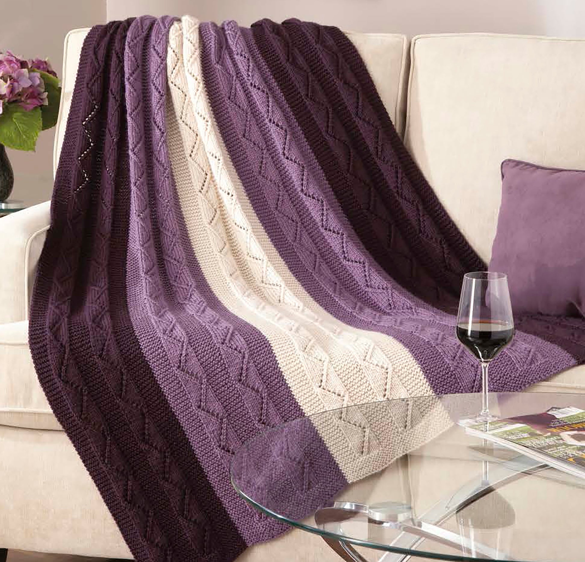 A beige sofa with a purple pillow is draped with the Decorator Throws—The Best of Mary Maxim by Leisure Arts, featuring purple and cream stripes. Nearby, a glass table holds red wine and a magazine, with flowers in the background.