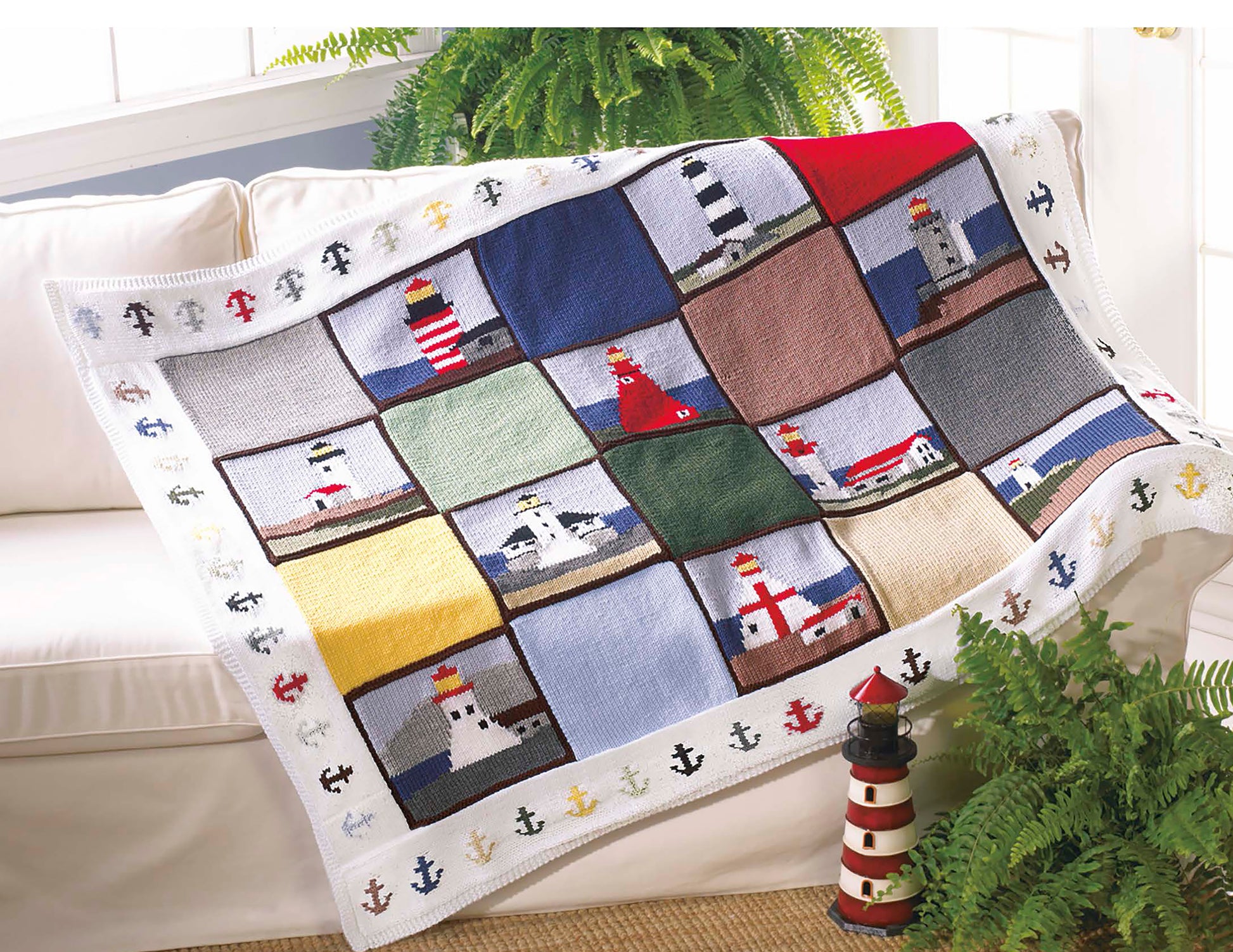The Leisure Arts "Decorator Throws—The Best of Mary Maxim" knit blanket features colorful lighthouse squares and anchor borders, adding cozy charm to a white sofa beside a green fern.