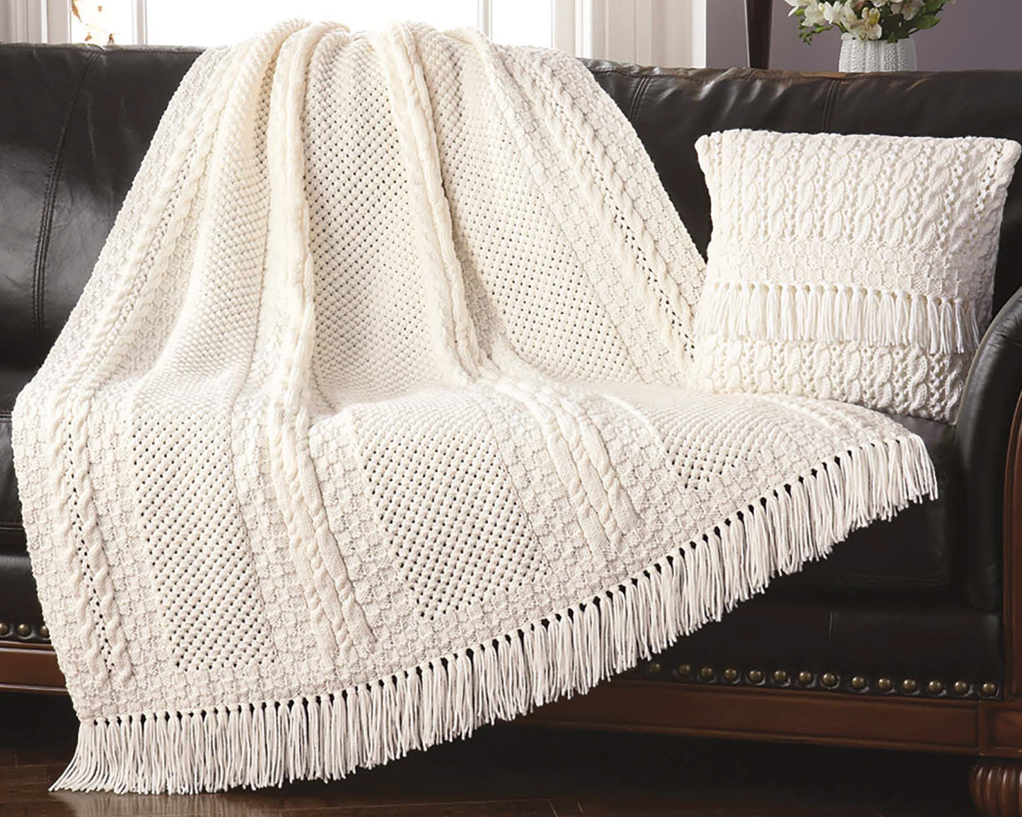 A textured white knit throw from "Decorator Throws—The Best of Mary Maxim" by Leisure Arts, with fringe, is draped over a dark leather sofa beside a matching pillow; in the background is a vase of flowers on a table.