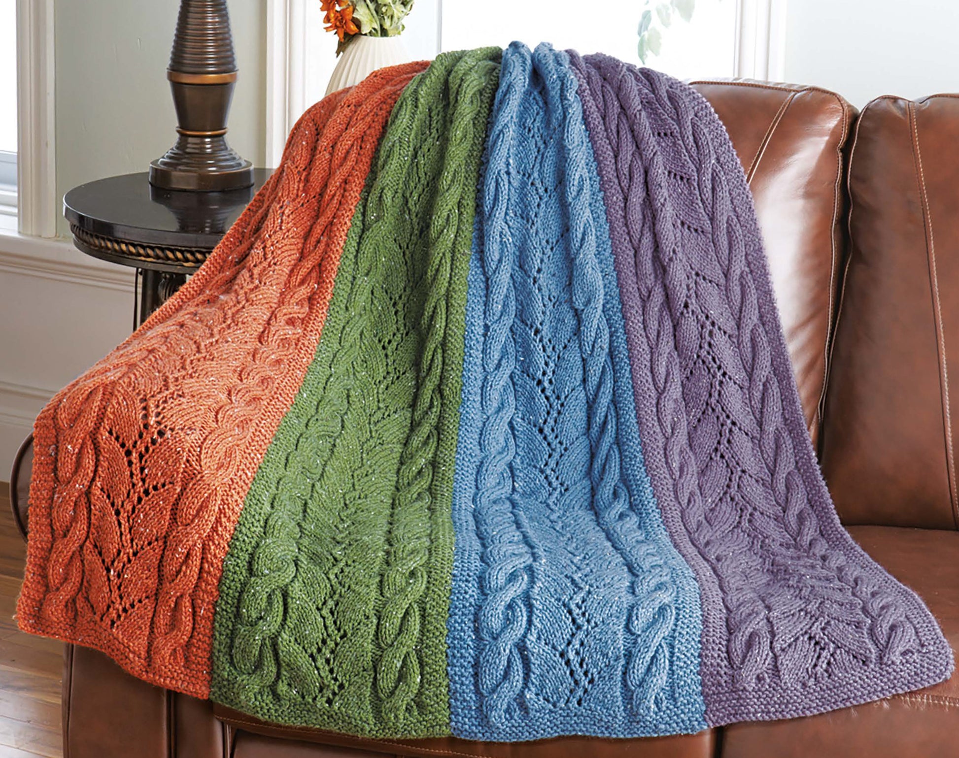 The Decorator Throws—The Best of Mary Maxim by Leisure Arts, a medium weight knitted blanket with orange, green, blue, and purple stripes plus cable and leaf patterns, is draped over a brown leather sofa in a cozy living room.