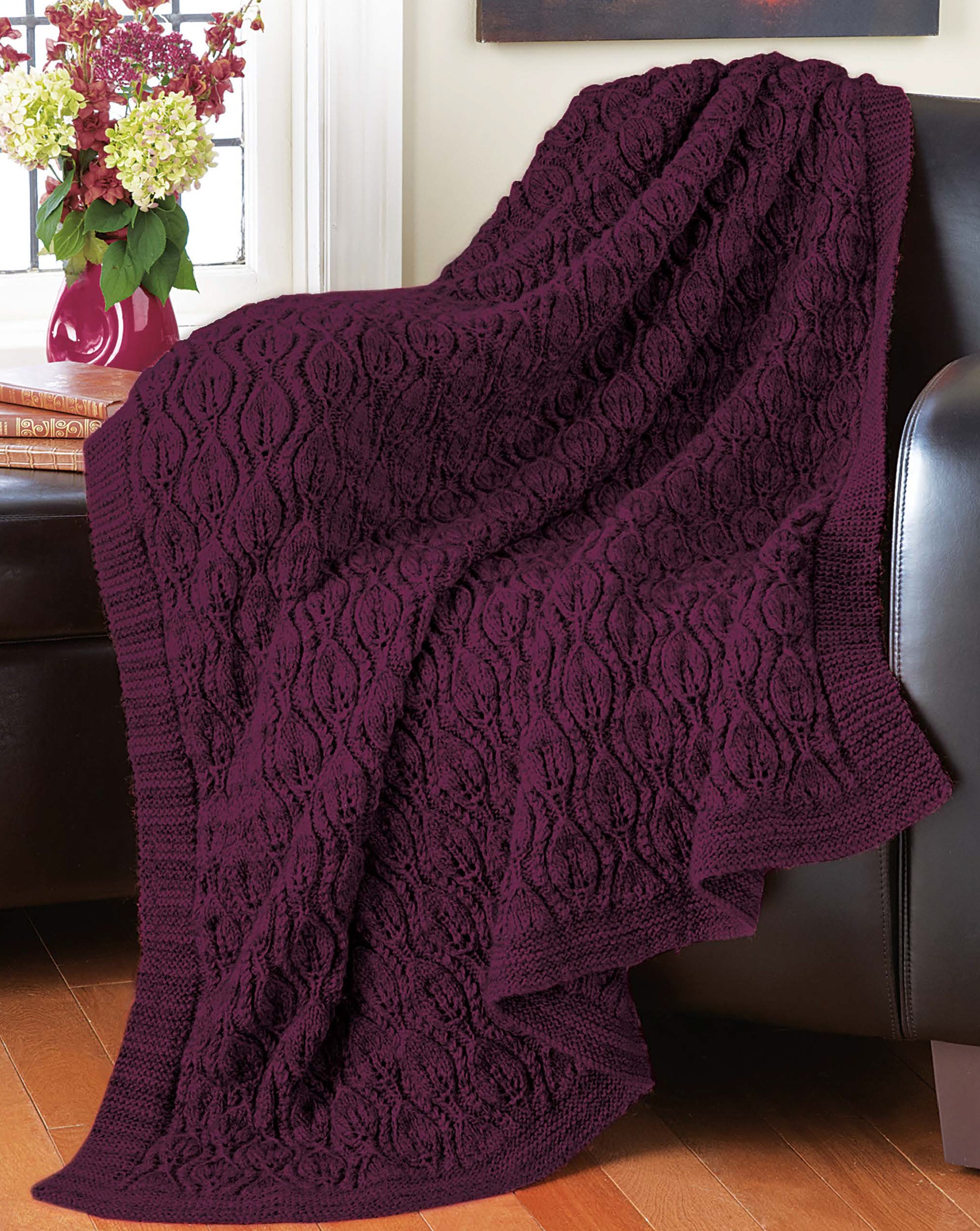 The Decorator Throws—The Best of Mary Maxim by Leisure Arts, a dark purple knit throw made from medium weight yarn, rests on a black leather sofa. Behind it, red and white flowers in a vase sit on a sunlit windowsill.