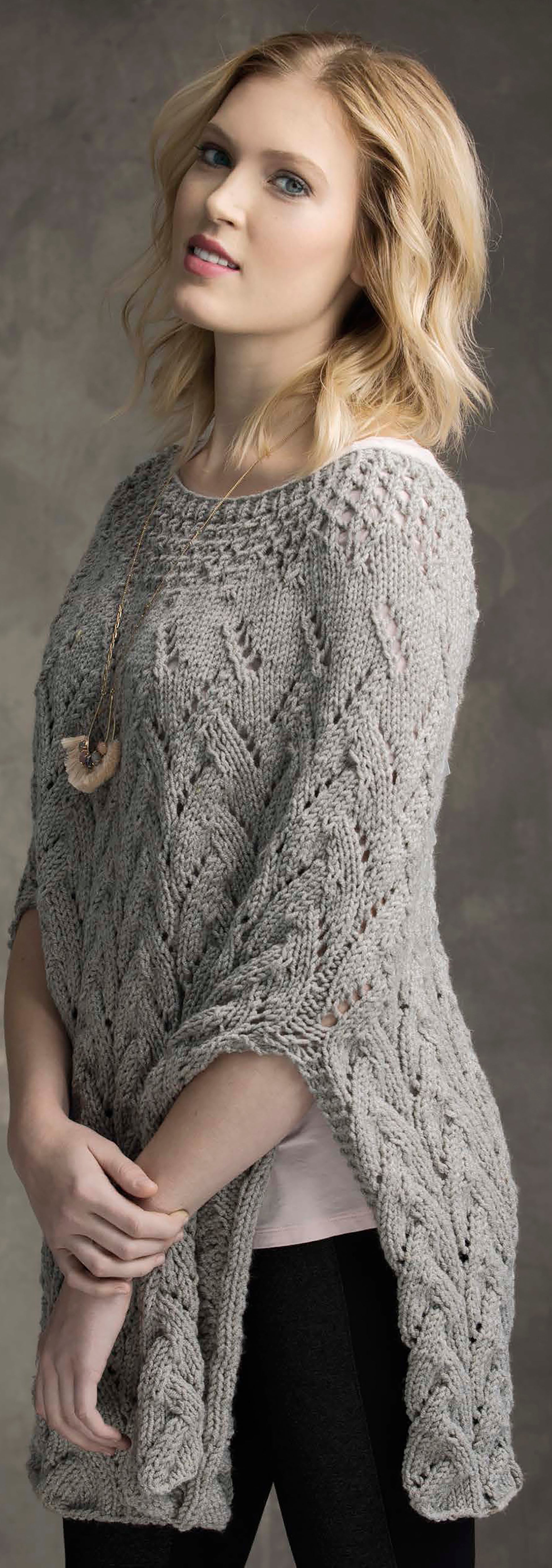 A woman with wavy blonde hair models a light gray knit sweater from Leisure Arts' "Wraps & Ponchos to Knit Digital Download," layering it over a pale top and dark pants, standing relaxed against a neutral background.