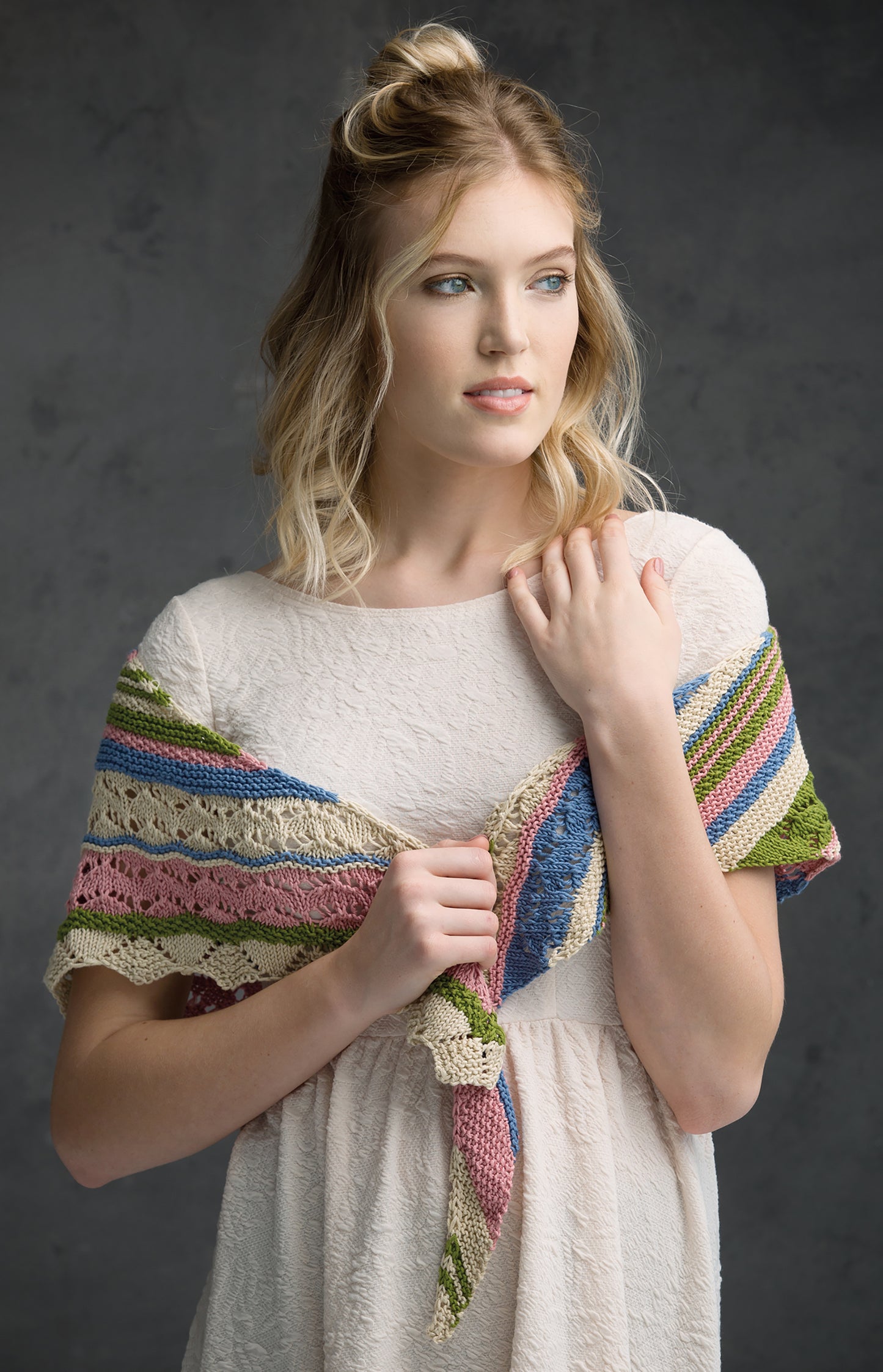 A young woman wears a textured white dress and a colorful knit wrap from Leisure Arts' "Wraps & Ponchos to Knit" digital download, draped around her shoulders against a dark background, highlighting beginner-friendly knitting charm.