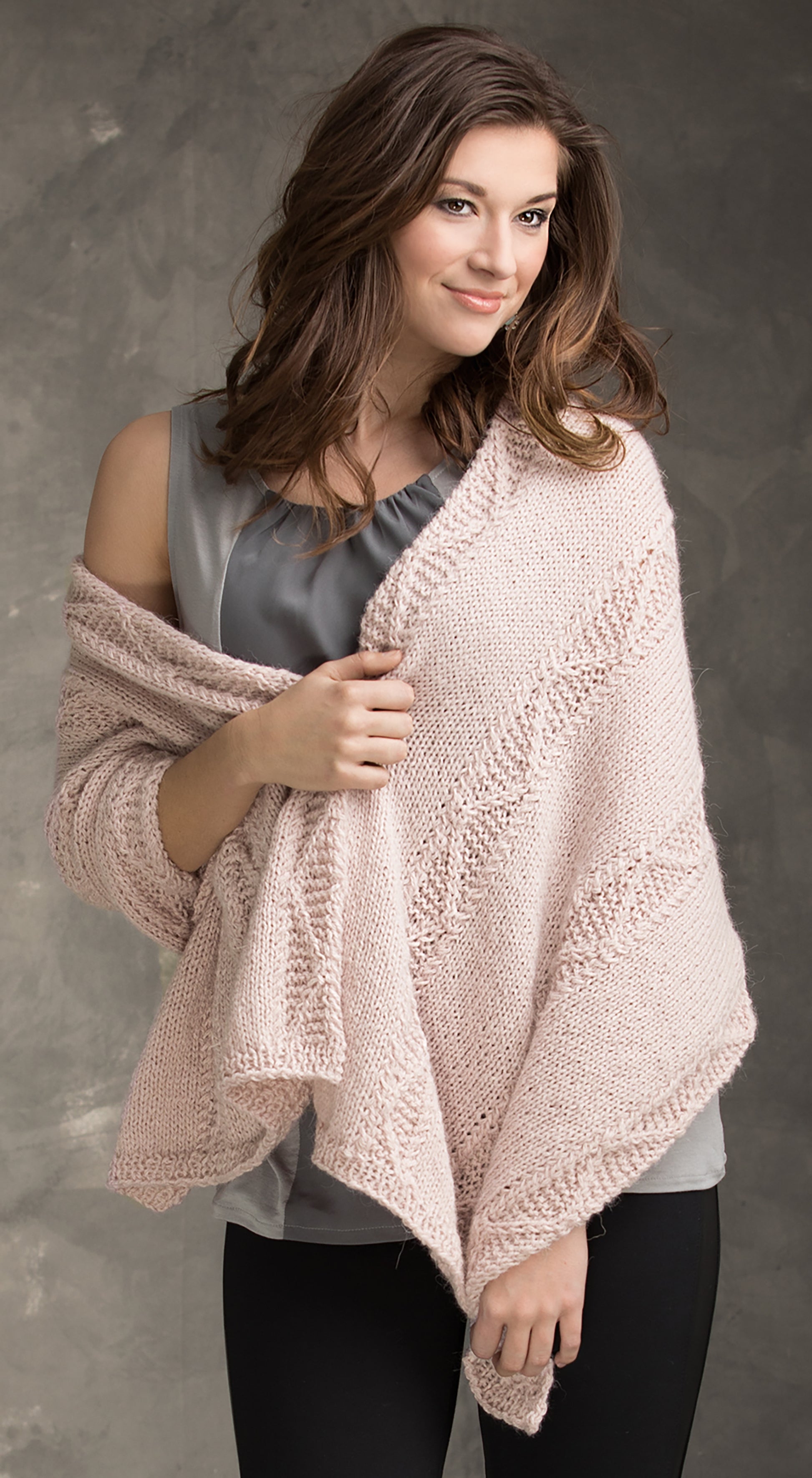 A woman with wavy brown hair models a light pink knitted shawl from the "Wraps & Ponchos to Knit" digital download by Leisure Arts, styled over a gray sleeveless top and black pants against a textured gray background, smiling softly.