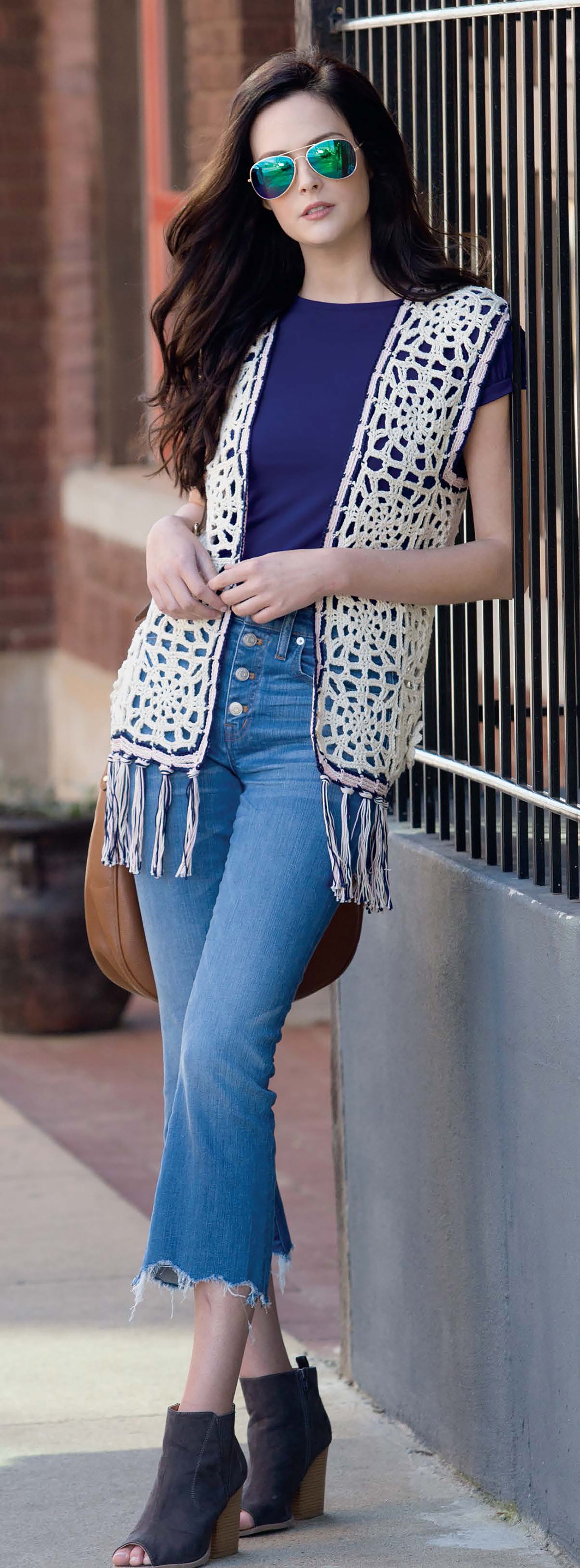 A woman in aviator sunglasses, navy top, and a cream fringe vest stands outdoors by a black fence—showcasing Leisure Arts’ “Crochet Vests The Long & Short of It,” perfect for beginners or anyone wanting trendy crochet vest patterns.