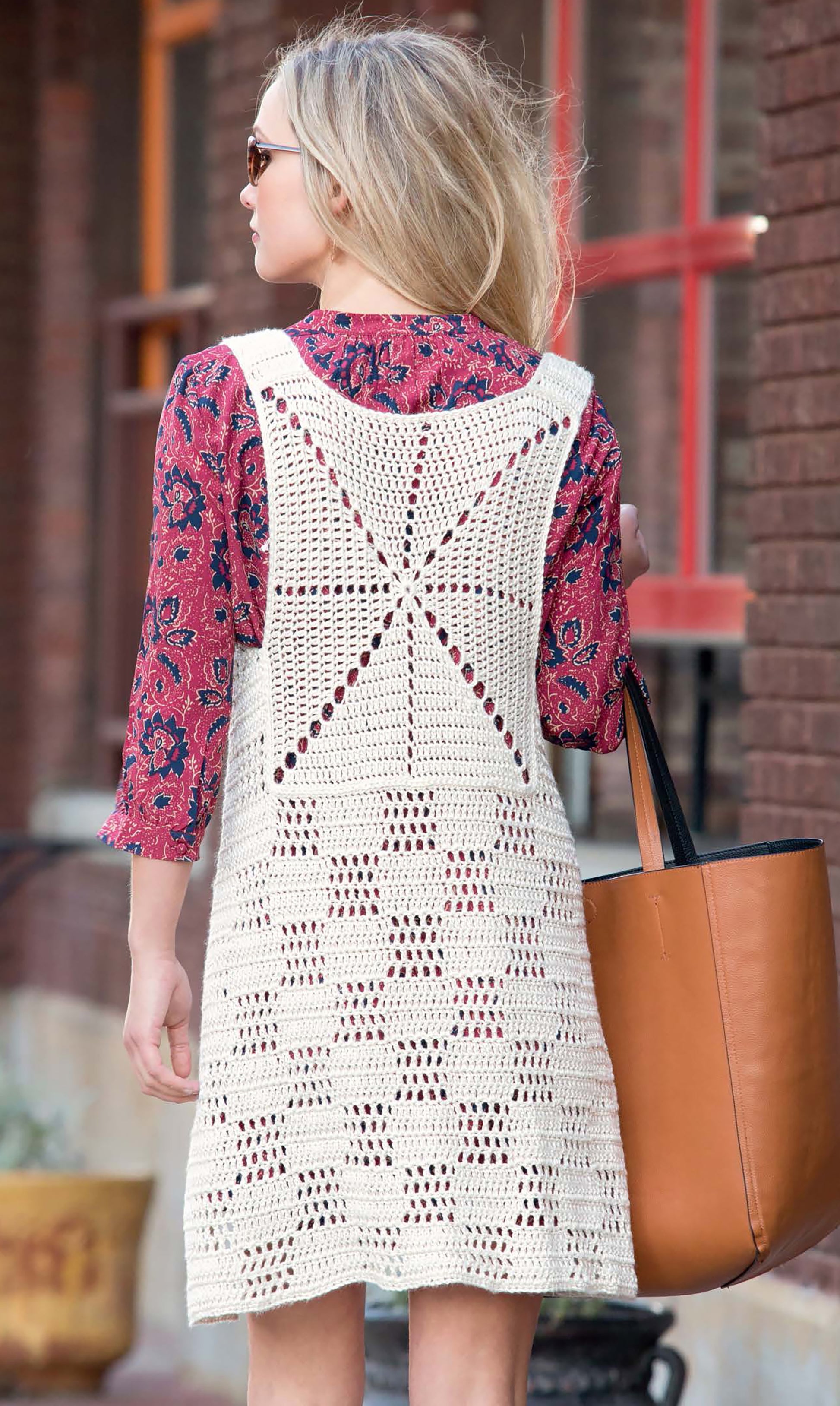 A woman with blonde hair and sunglasses walks outside in a red patterned top, wearing the Leisure Arts "Crochet Vests The Long & Short of It," and carries a large tan tote bag. Perfect for beginners or those exploring crochet vest patterns.