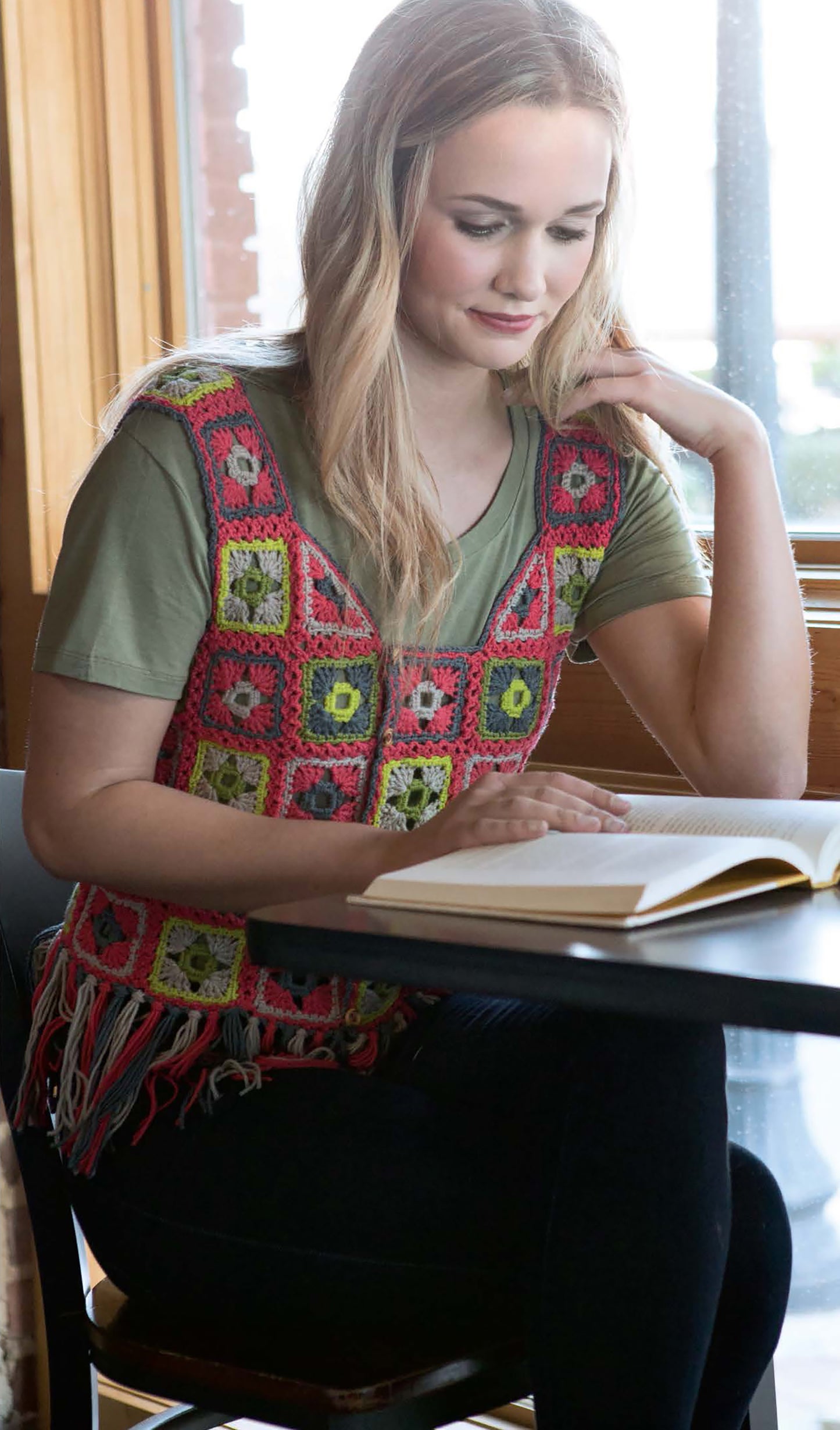 A woman with long blonde hair reads by a window, wearing a green t-shirt and the colorful Crochet Vests The Long & Short of It by Leisure Arts, featuring red, pink, and yellow squares with fringe at the bottom.