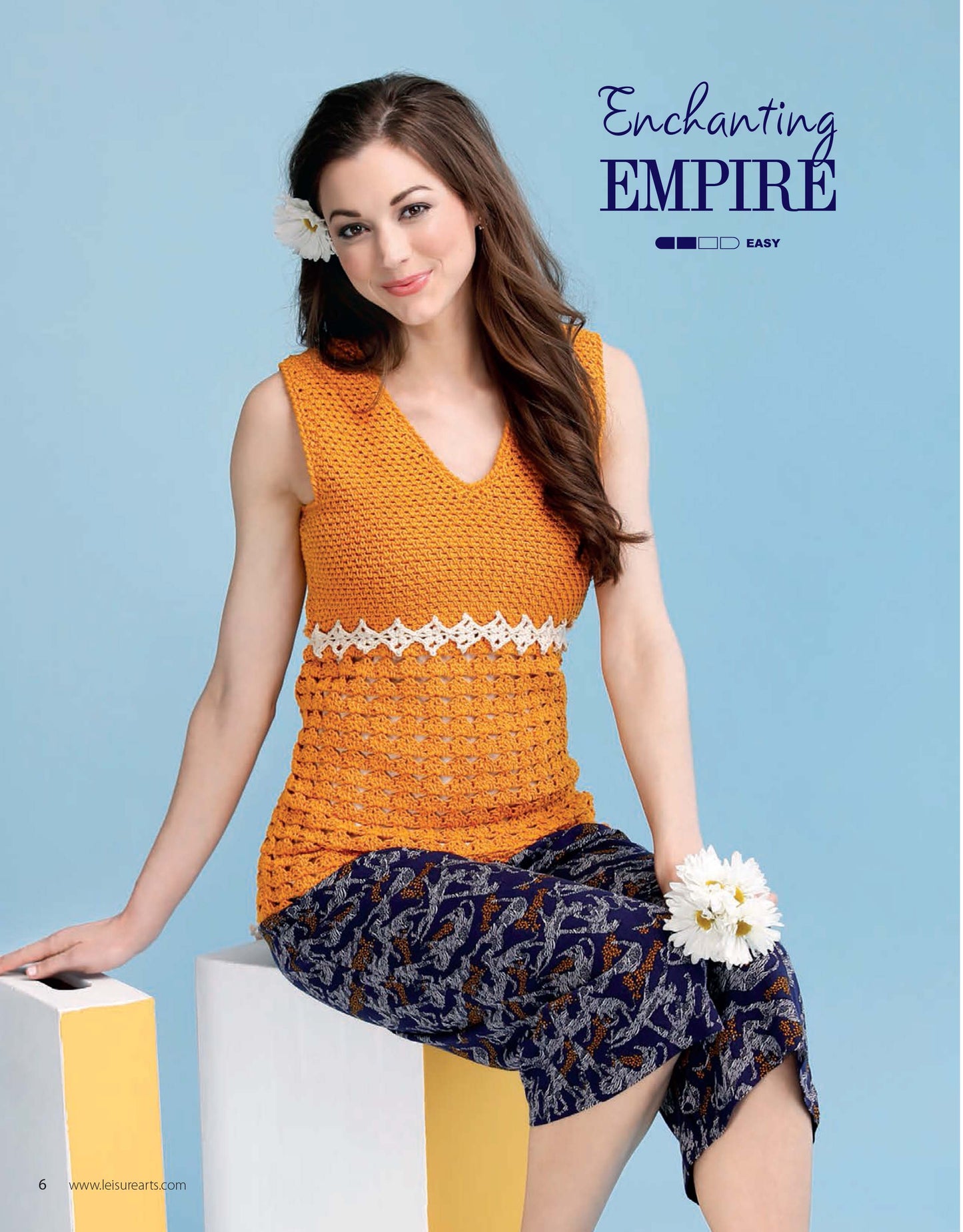 A woman with long brown hair models an orange sleeveless top from Leisure Arts' "Crochet Super Simple Summer Tops," paired with a blue patterned skirt, sitting on white blocks and holding white flowers against a light blue backdrop.