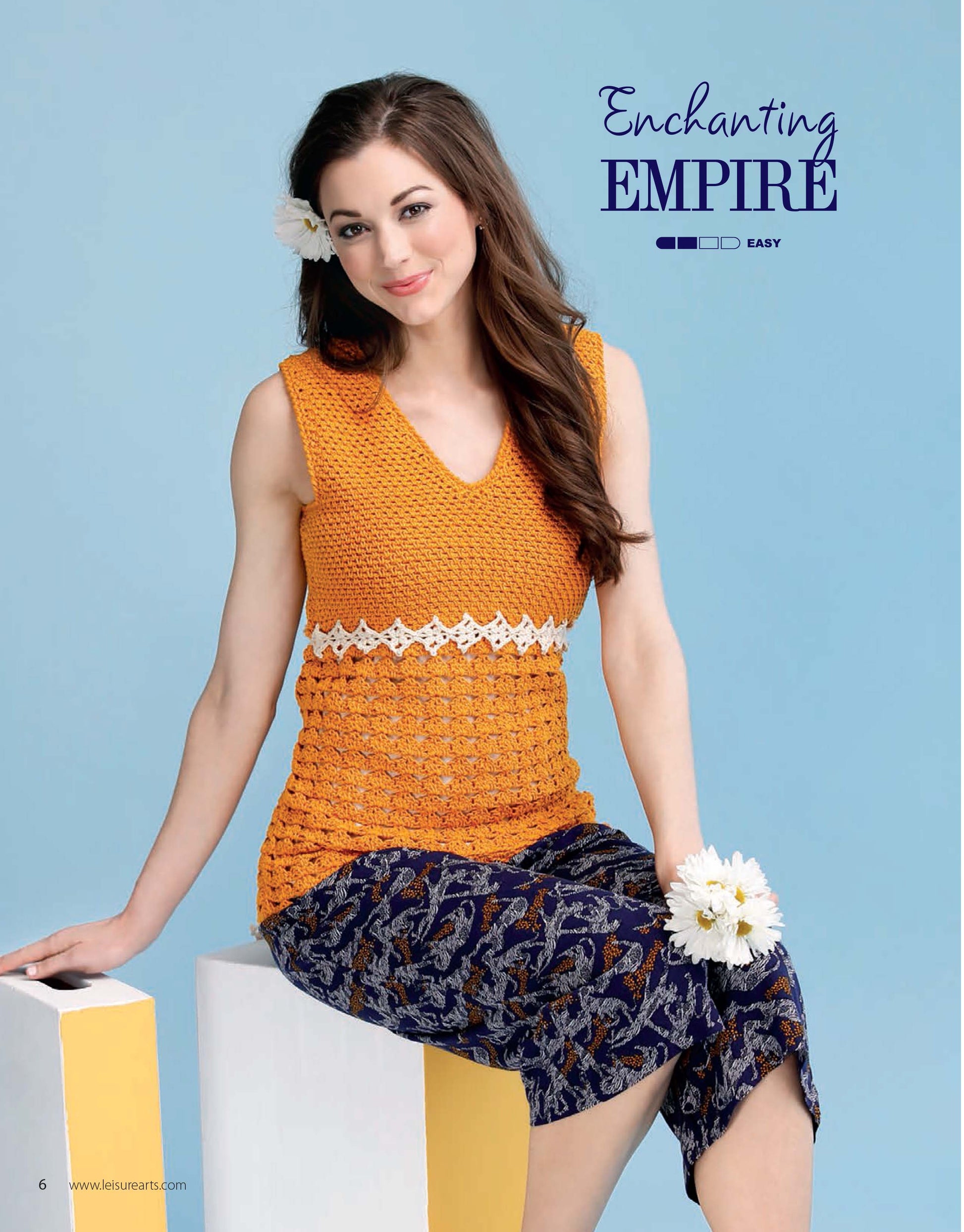 A woman with long brown hair models an orange sleeveless top from Leisure Arts' "Crochet Super Simple Summer Tops," paired with a blue patterned skirt, sitting on white blocks and holding white flowers against a light blue backdrop.