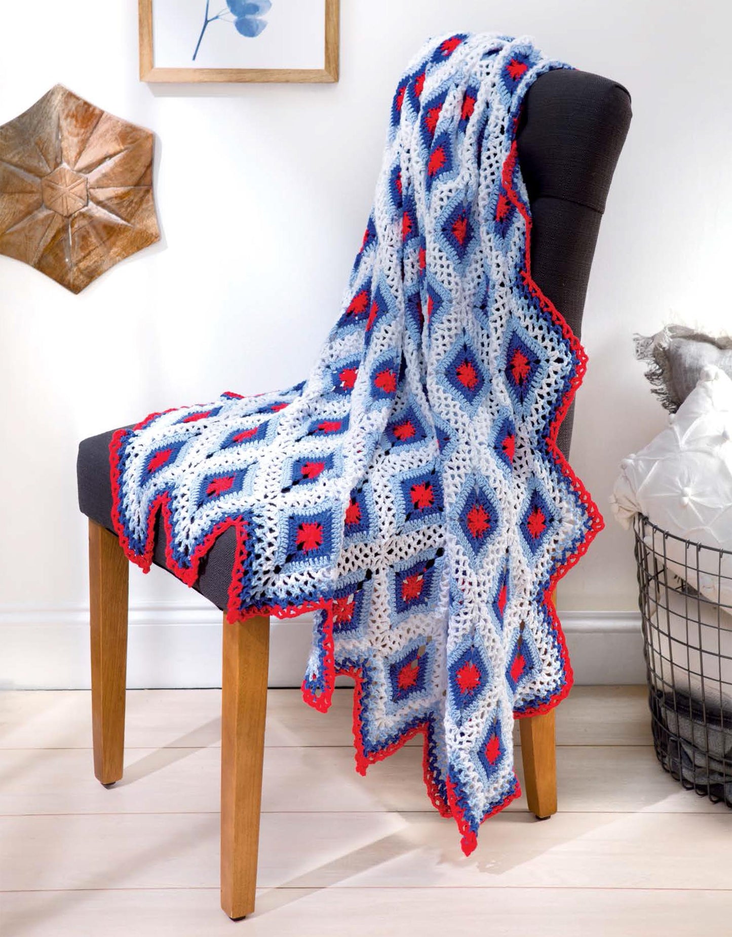 A Leisure Arts Cotton Blanket & Throw with a blue, white, and red diamond pattern is draped over a dark upholstered chair in a bright room with wooden floors and wall art.