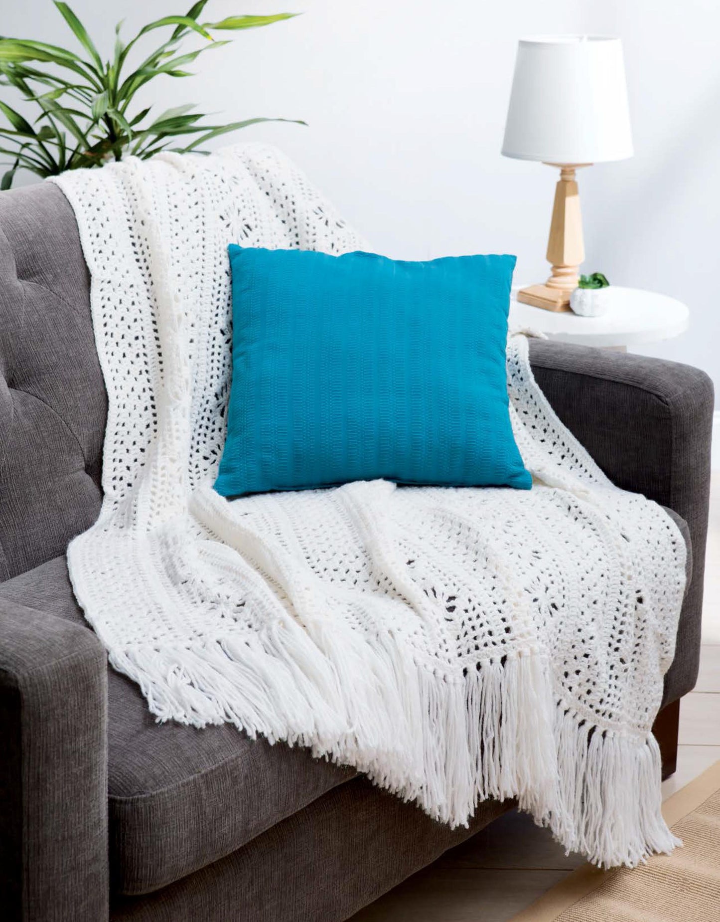 A grey sofa features a bright blue pillow and a Leisure Arts Cotton Blanket & Throw in white crochet, draped stylishly. In the background, a wooden side table holds a white lamp and small plants.