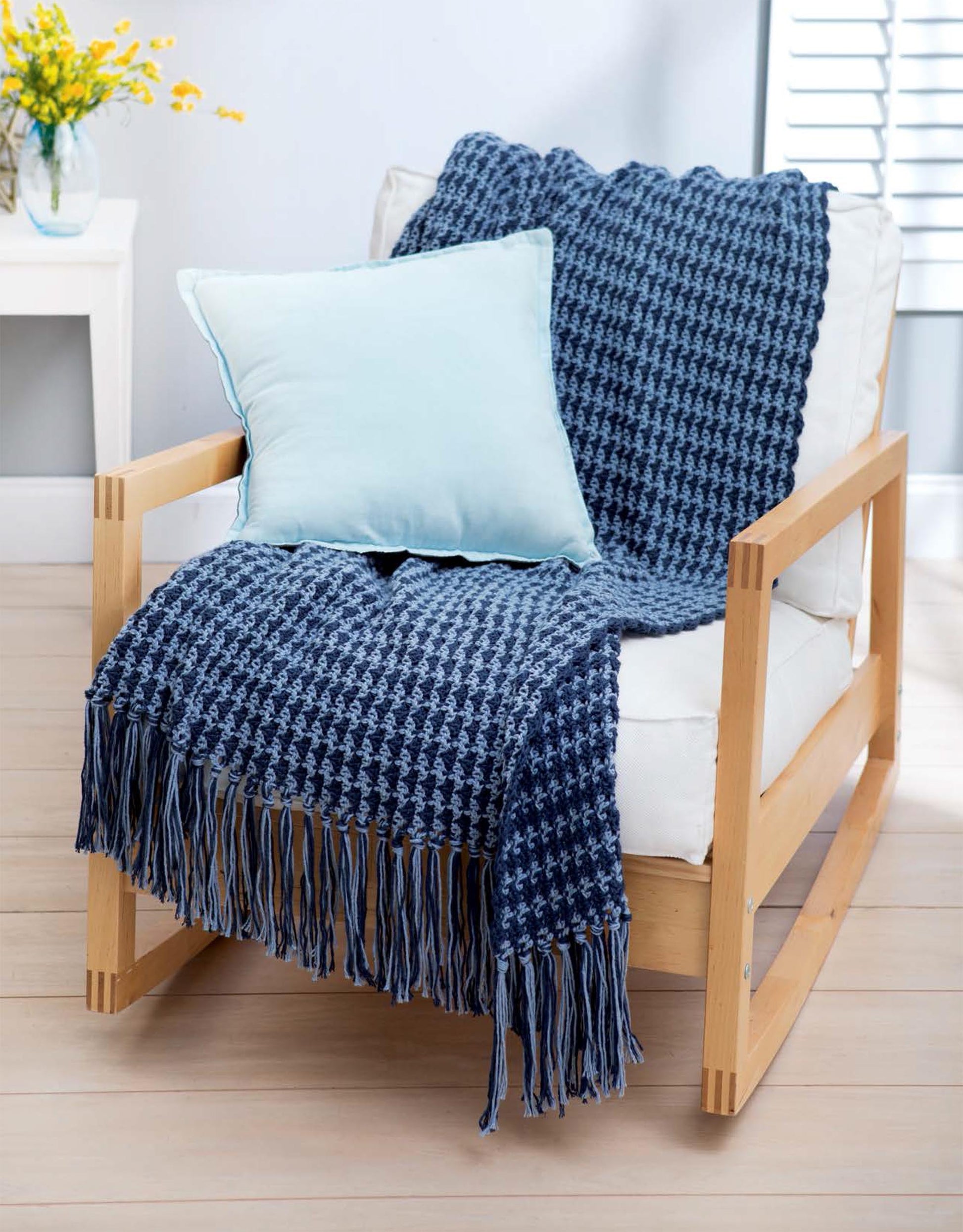 A cozy wooden armchair features a white cushion, a Leisure Arts Cotton Blanket & Throw in blue medium weight yarn, and a light blue pillow. In the background, a side table holds a vase of yellow flowers.