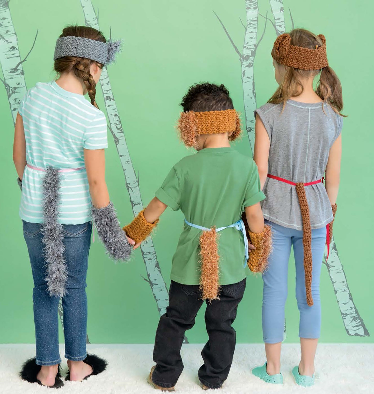 Three kids in crochet animal costumes—furry tails, ear headbands, and mittens—hold hands against a tree wall. Featured: Crochet Kids Dress Up Digital Download by Leisure Arts, perfect for fun dress-up adventures.