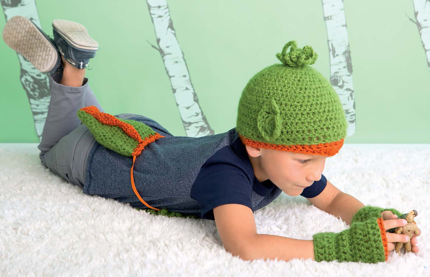 A child wears a green knot hat and fingerless gloves from the “Crochet Kids Dress Up Digital Download” by Leisure Arts—ideal beginner crochet costumes—playing on a white carpet with birch tree wall art in the background.