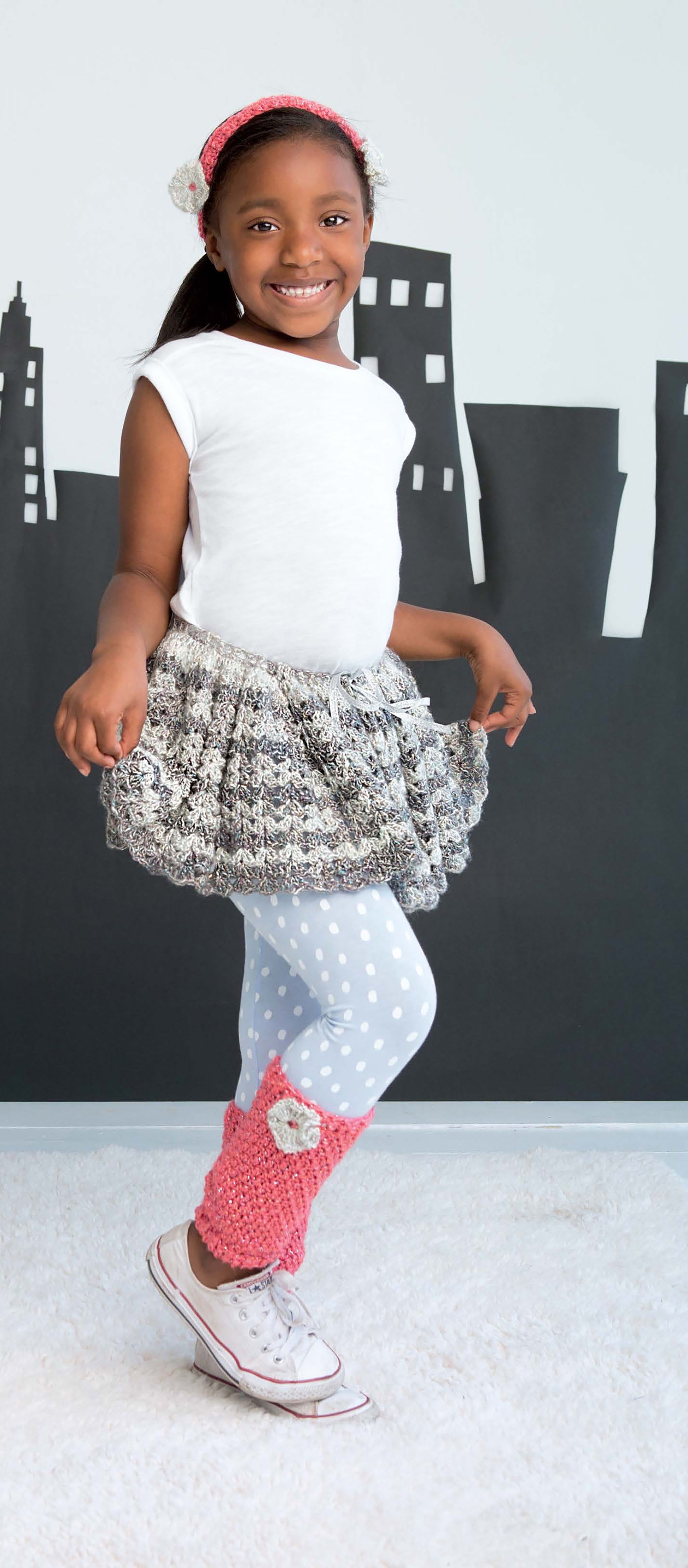 A smiling young girl poses before a cityscape, wearing pink crochet leg warmers made with Leisure Arts’ Crochet Kids Dress Up Digital Download—ideal for beginners—plus a white shirt, patterned skirt, polka-dot leggings, sneakers, and a cute pink headband.