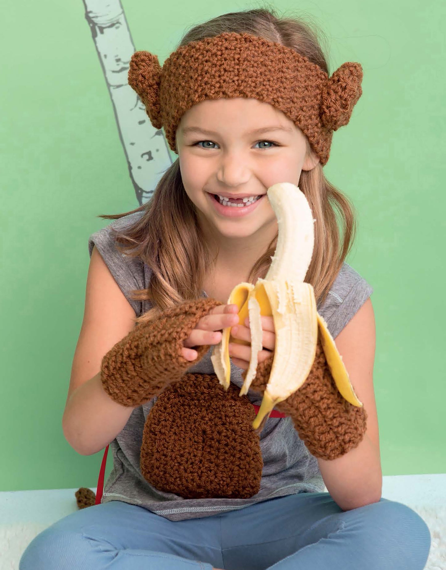 A smiling young girl wears the Leisure Arts Crochet Kids Dress Up Digital Download monkey headband, gloves, and chest piece, holding a peeled banana against a green backdrop with tree illustration—ideal for crochet costumes and creative play.