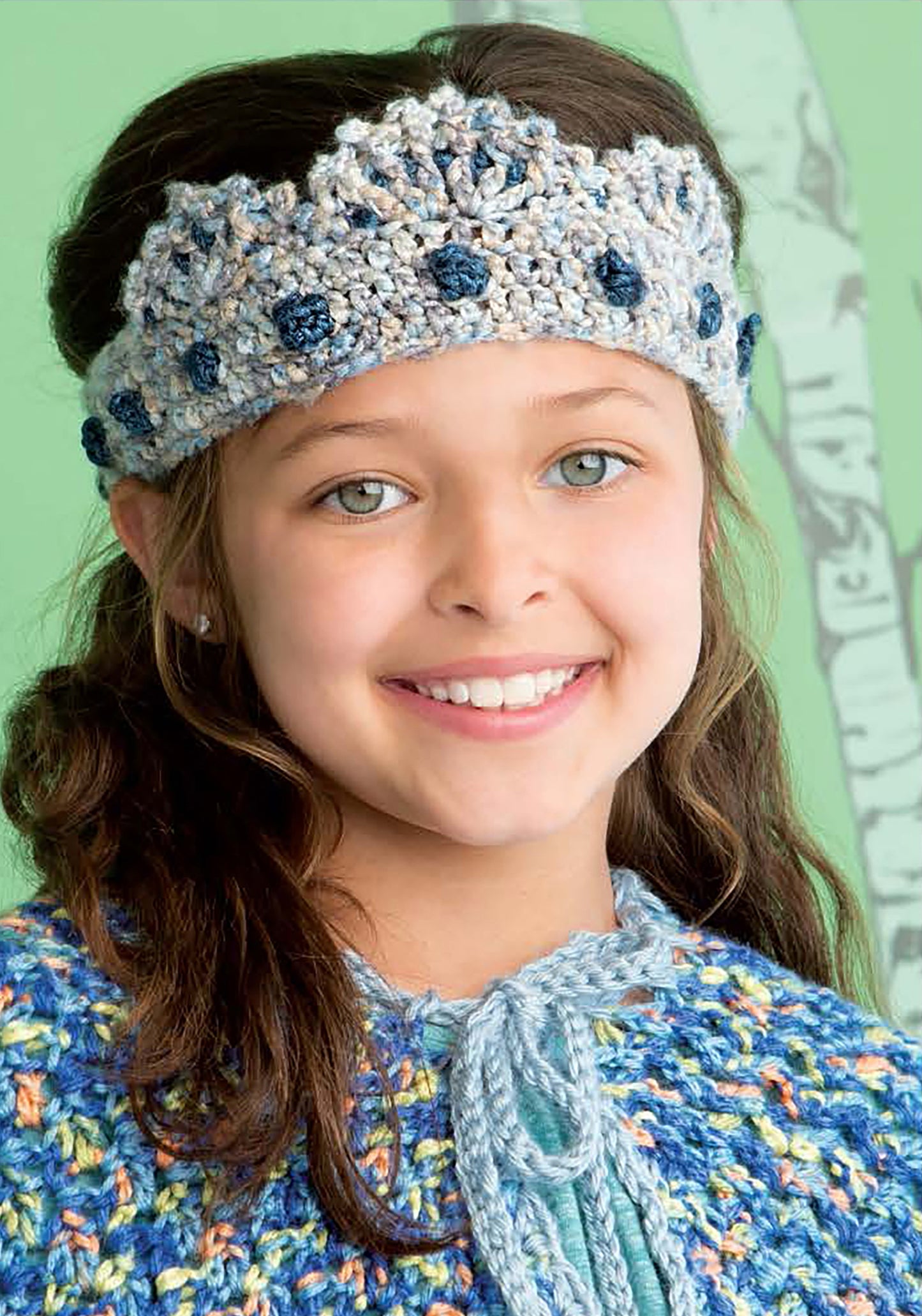A smiling young girl with light eyes and brown hair wears a blue and gray crown headband from the Crochet Kids Dress Up Digital Download by Leisure Arts, standing before a green background with a tree illustration.