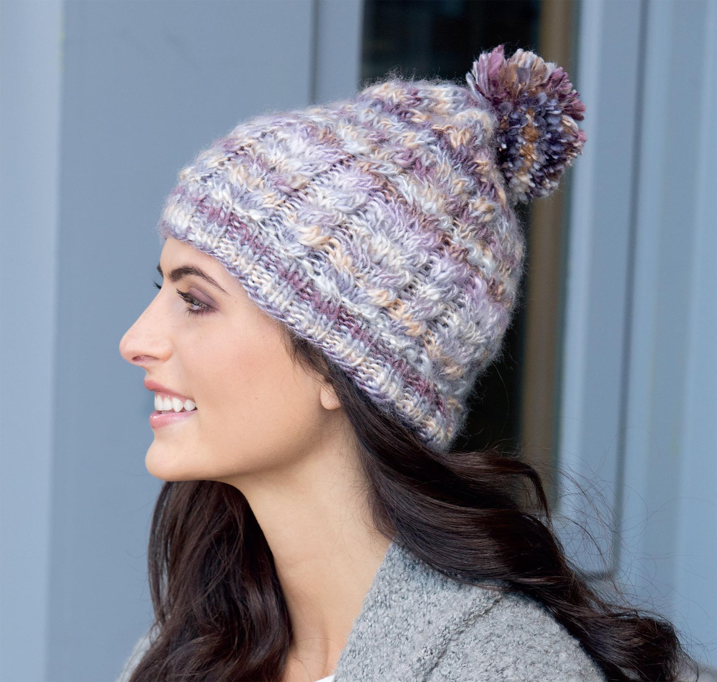 A woman with long dark hair smiles while wearing a Leisure Arts "Hats for the Family" knit beanie in purple, gray, and cream—a cozy accessory ideal for showcasing beginner projects or experimenting with new knitting techniques.