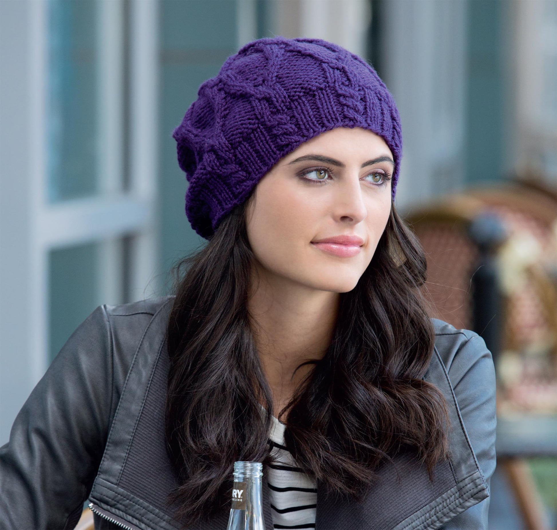A woman with long dark hair sits outdoors at a table, looking thoughtful. She wears a purple beanie made from Leisure Arts' "Hats for the Family," along with a striped shirt and black leather jacket.