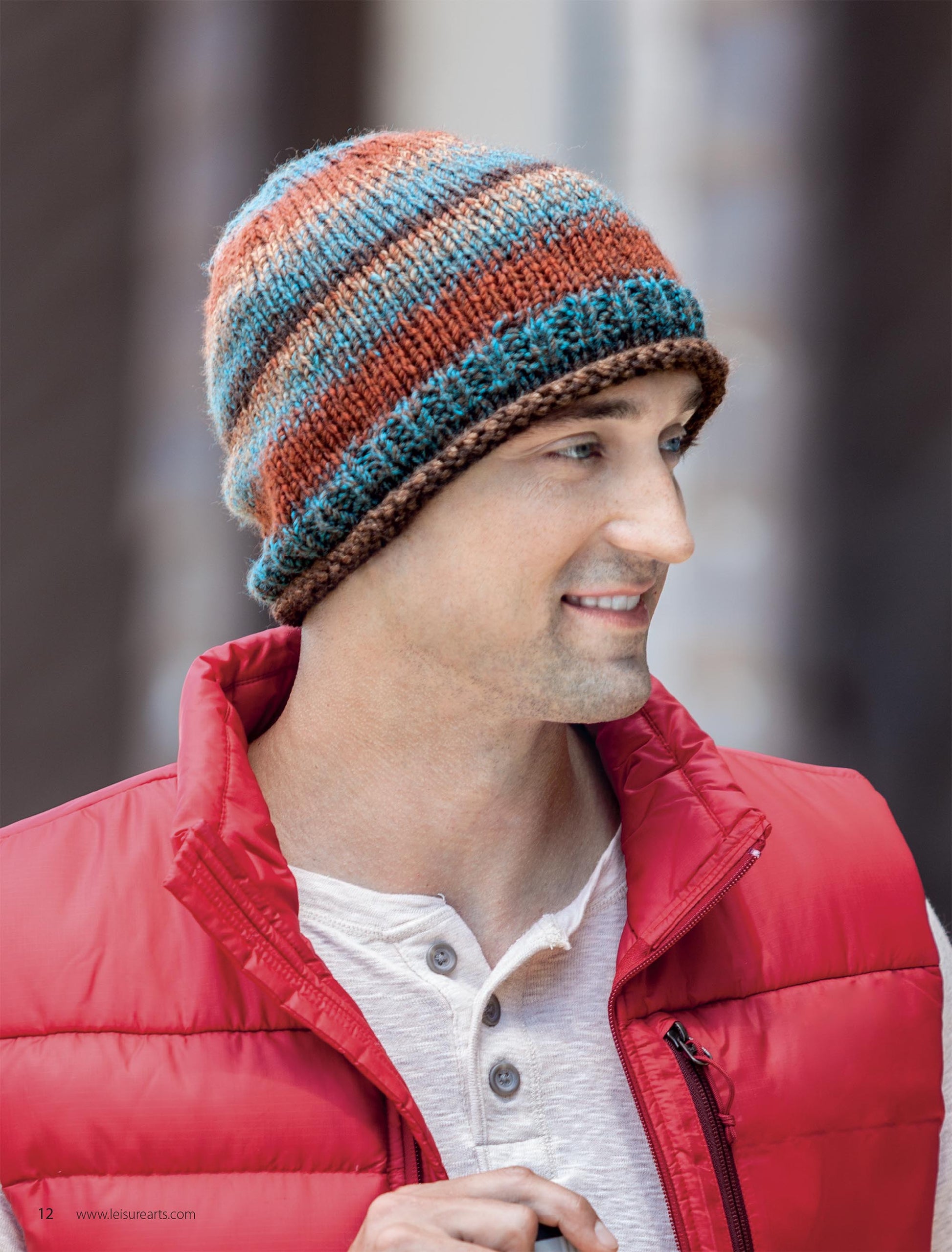 A man in a red puffer vest and cream henley smiles in an urban setting, modeling a blue, brown, and rust-striped knit beanie from Leisure Arts' "Hats for the Family," a perfect project idea for beginner knitters.