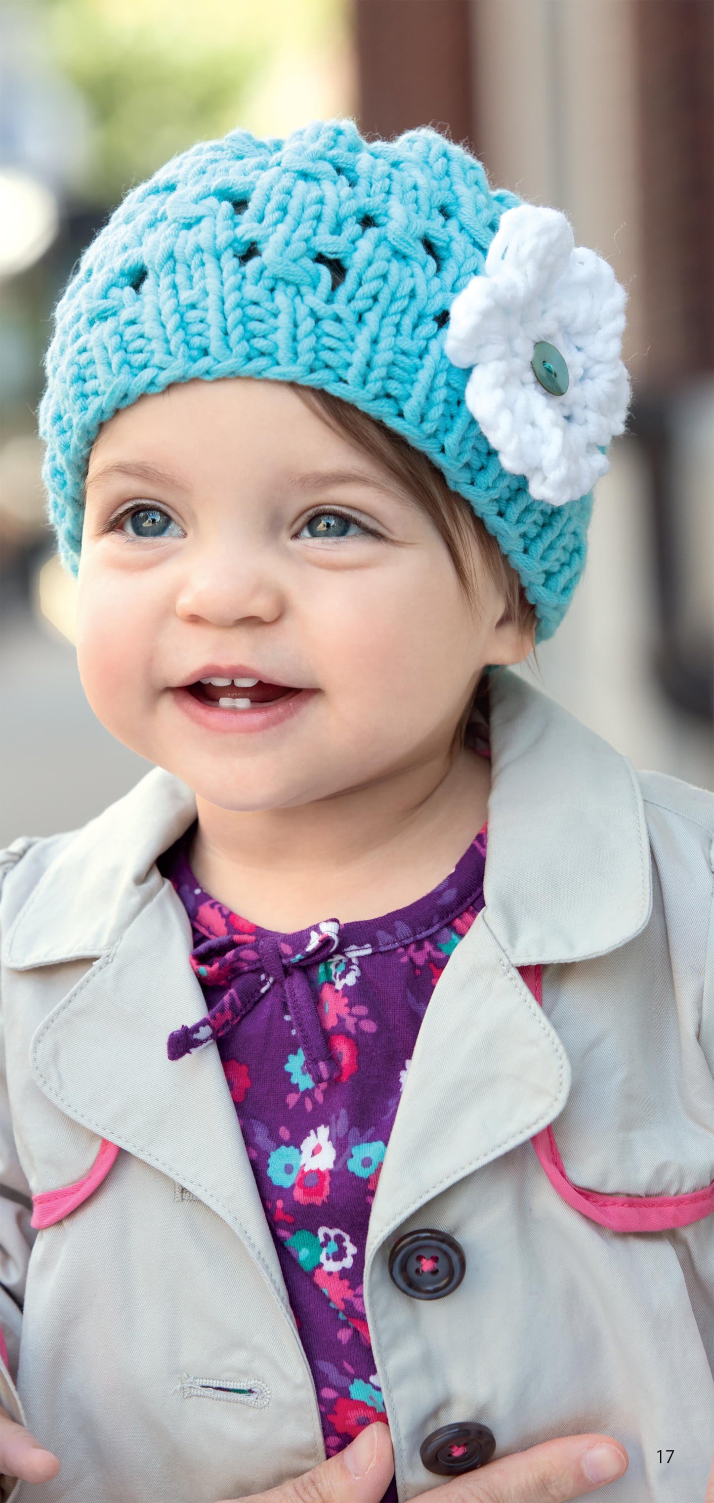 A smiling toddler with blue eyes wears a turquoise knit hat from Leisure Arts’ Hats for the Family, featuring a white flower, paired with a light coat over a purple floral dress while standing outdoors.