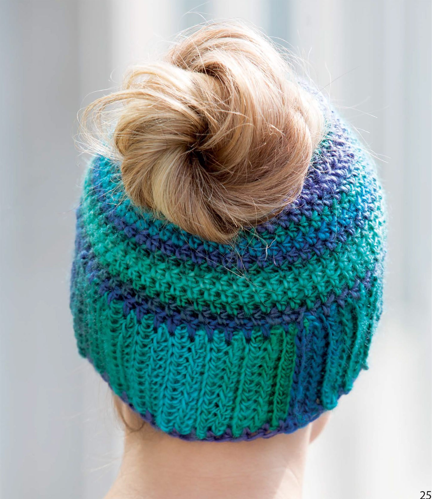 A person with blonde hair in a messy bun wears the Leisure Arts Messy Bun Hats, Plus!—a blue and green knit slouchy hat with an opening on top for their bun—set against a softly blurred background.
