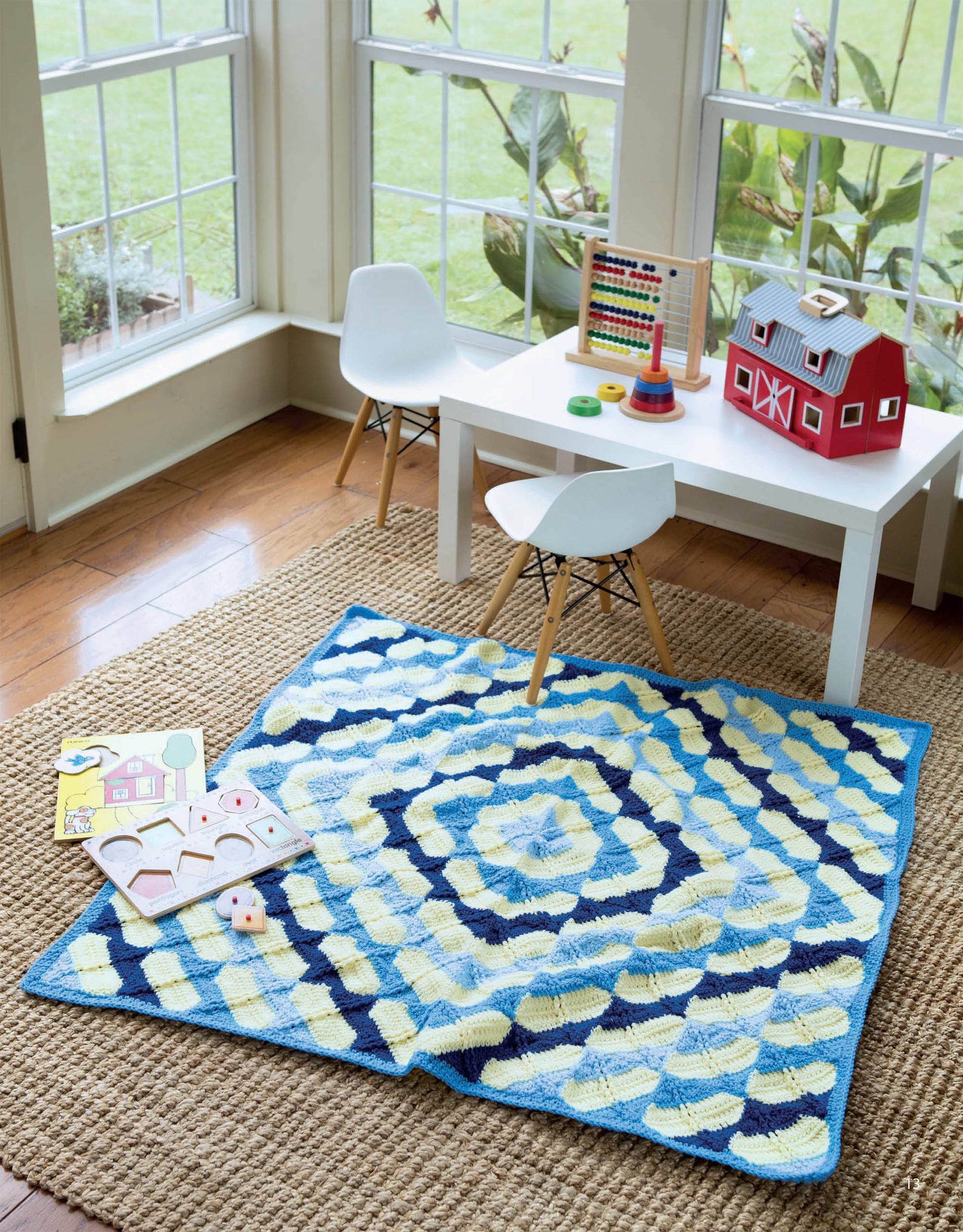A bright playroom with large windows, a white kids table and chairs, toys, and the Leisure Arts Baby Bright Quilts—Use Pops of Color in Stunning Baby Designs!—in blue, yellow, and cream displayed on a woven rug.